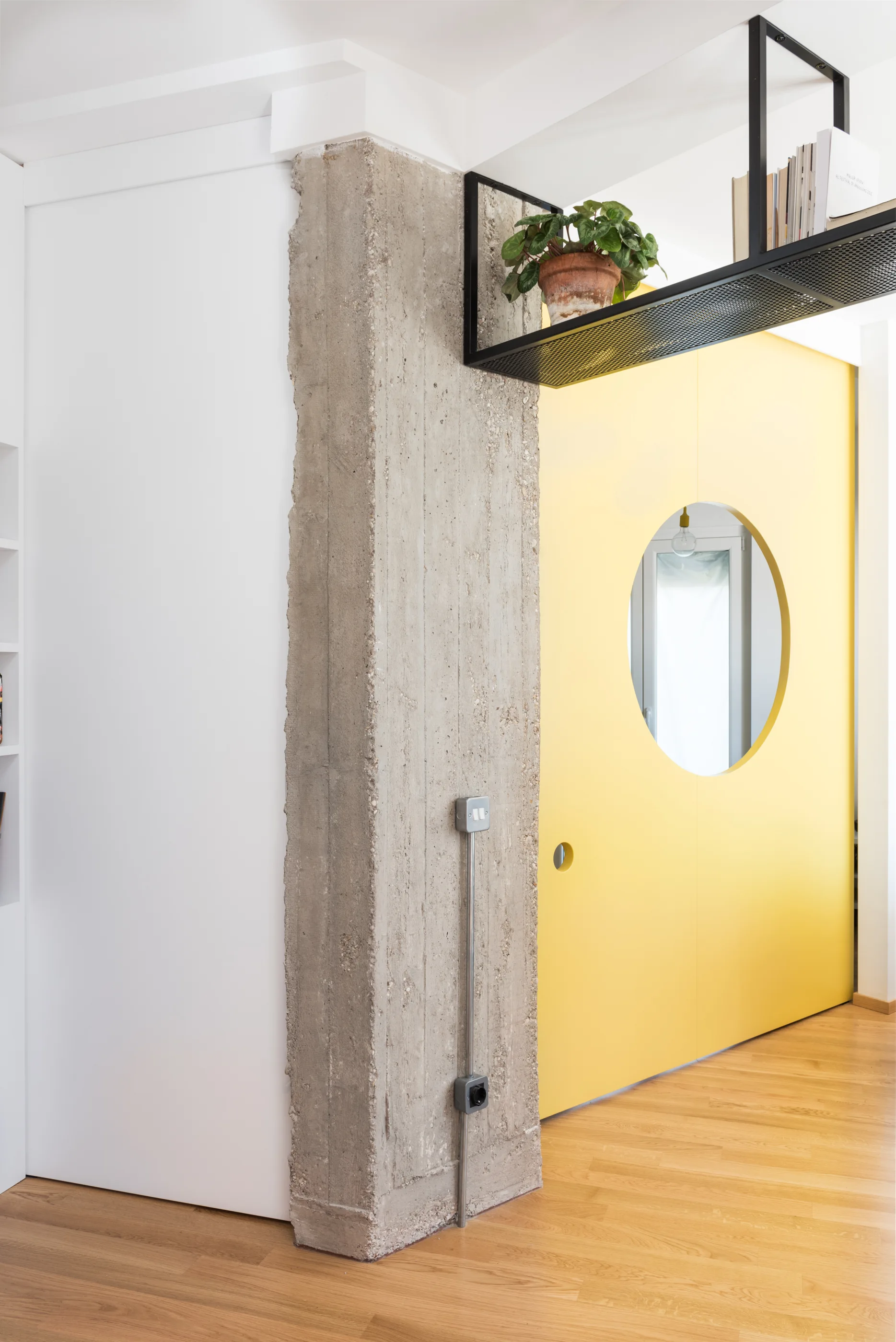 A bright yellow door with a circular cutout sits next to an exposed raw concrete pillar and below a black metal mesh overhead shelf.