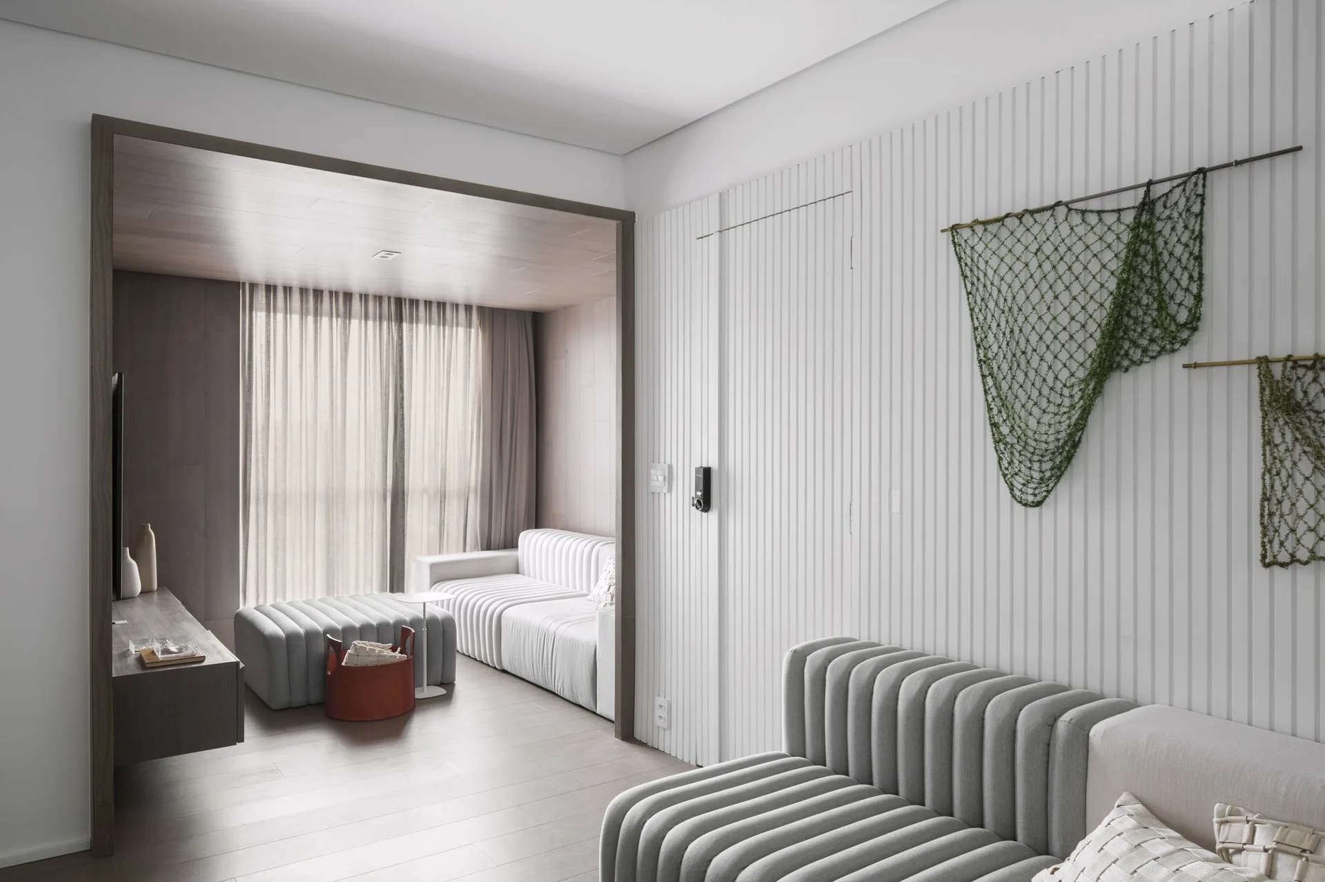 A contemporary living space with white vertical slatted wall paneling, grey ribbed sofas, a wooden room transition archway, and sheer curtains.