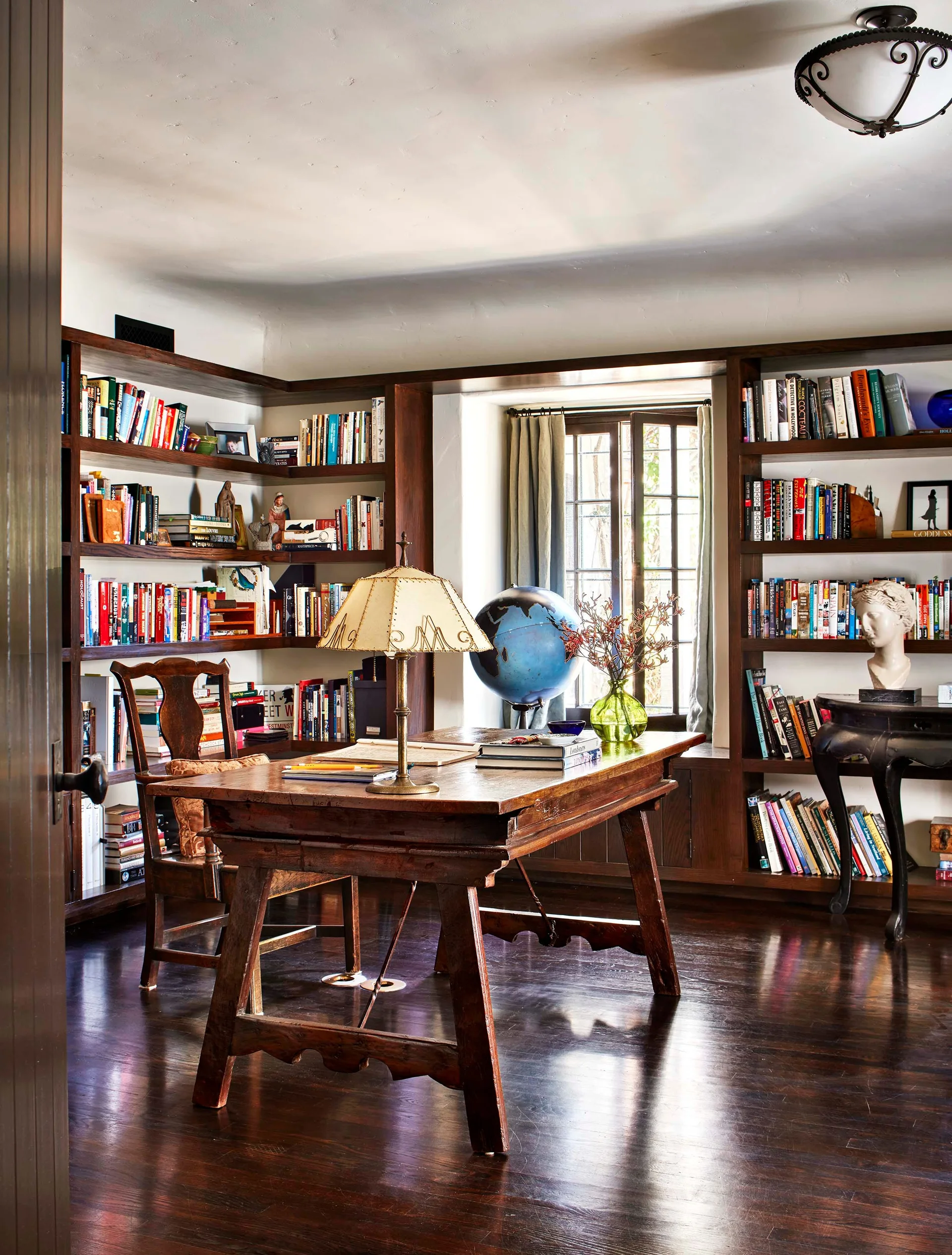 A traditional home office features a dark wood trestle desk, floor-to-ceiling built-in bookshelves filled with books, and polished hardwood floors.
