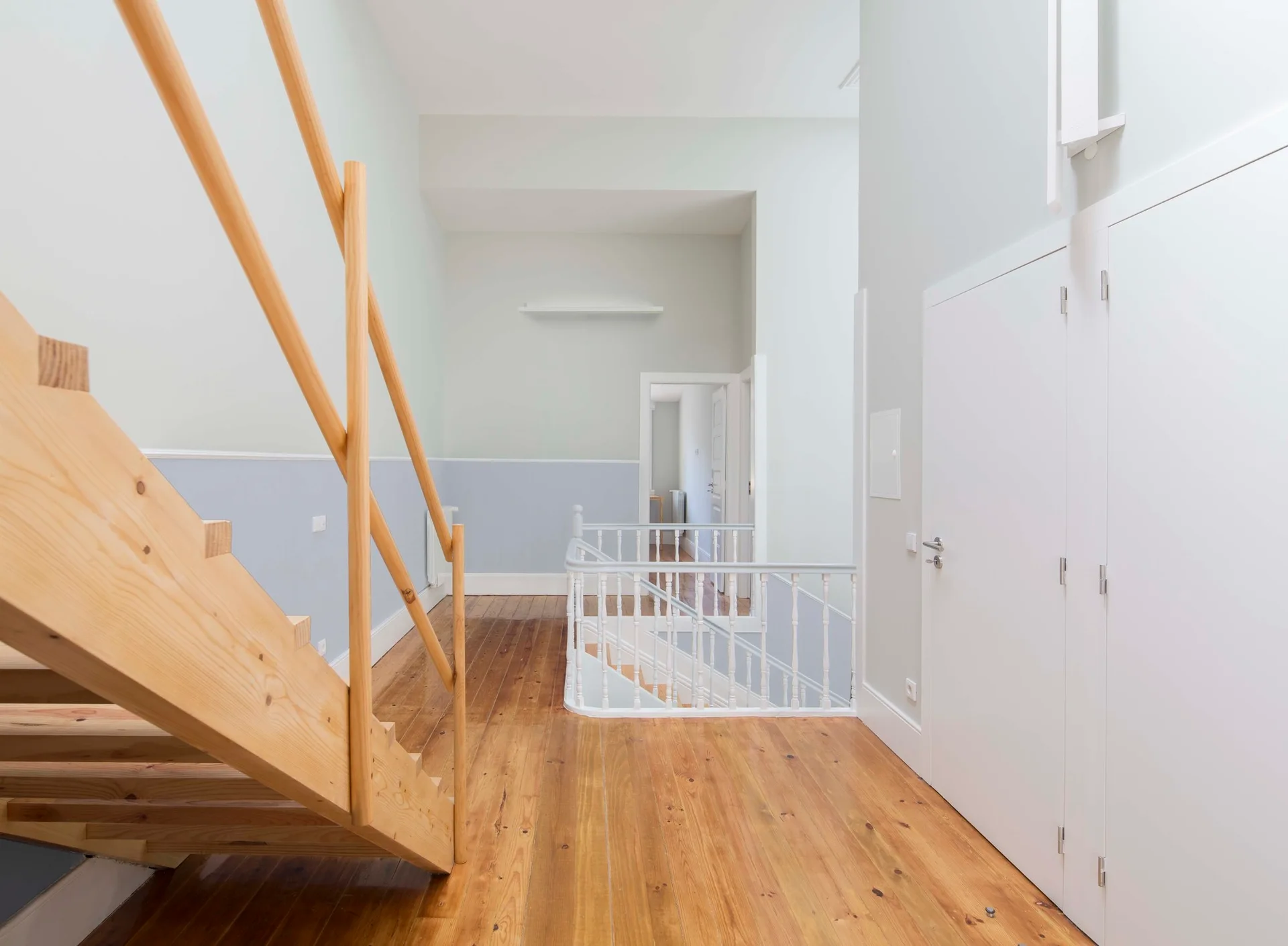 A bright upstairs landing features a natural wood staircase on the left, polished wood floors, and a white balustrade overlooking the lower level.