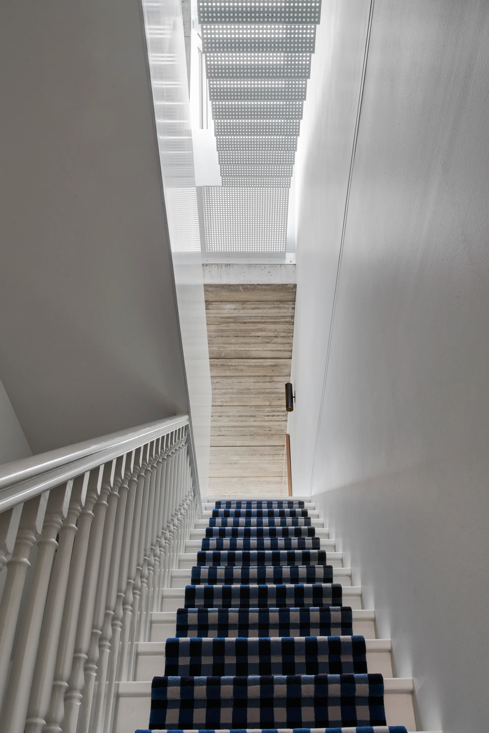 A vertical view looking up a white wooden staircase with a blue and black plaid runner toward a perforated white metal ceiling panel.