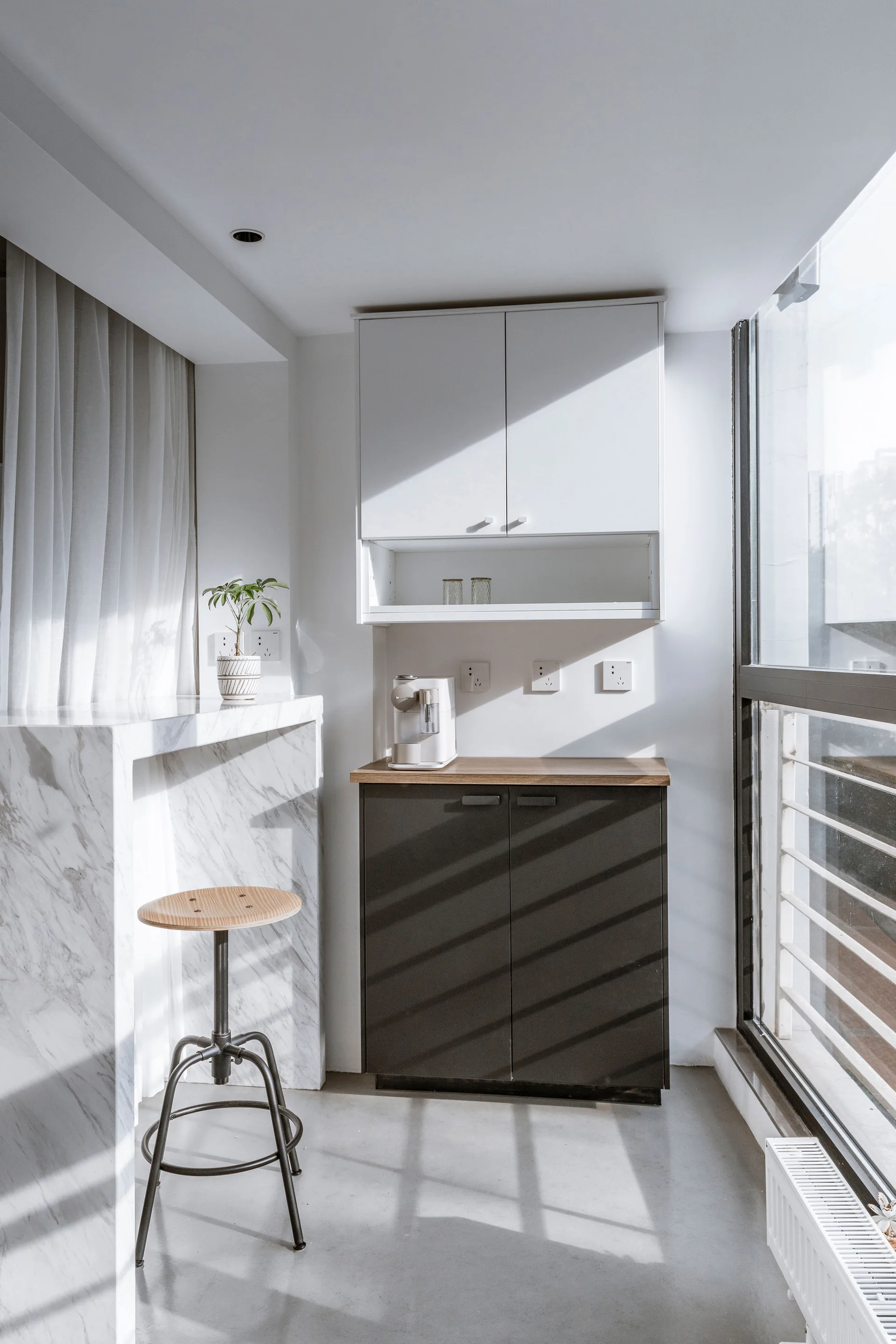 A minimalist kitchen nook features a veined marble waterfall counter, dark cabinets, and a wooden stool in bright morning sunlight.