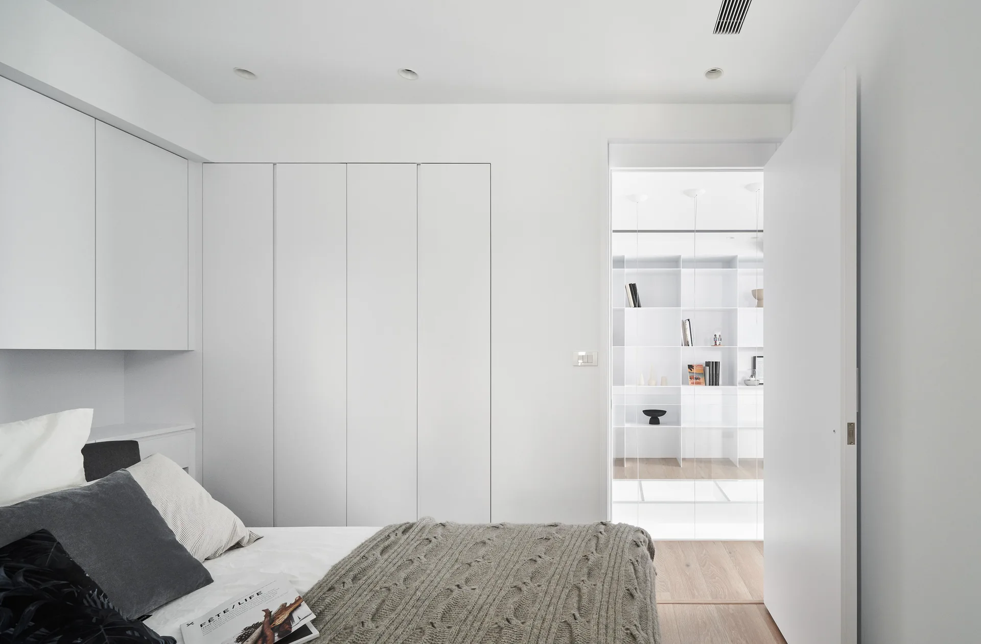 A minimalist bedroom features seamless white wardrobes, a bed with a grey cable-knit throw, and an open door showing a shelved closet beyond.