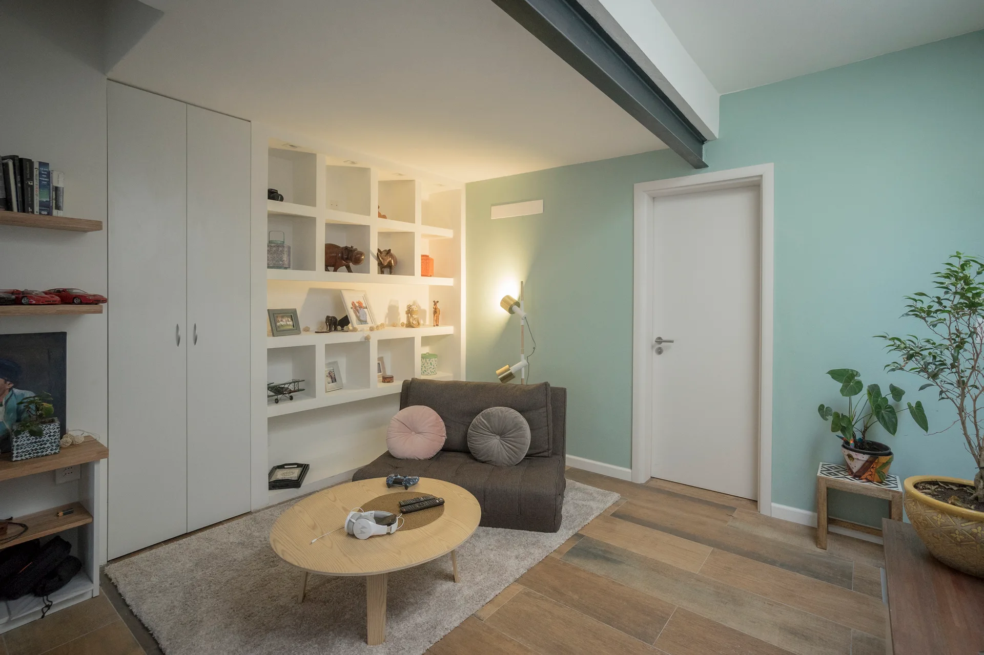 A modern living room features a dark grey floor sofa, a round light wood coffee table, mint green walls, and a recessed white grid shelving unit.