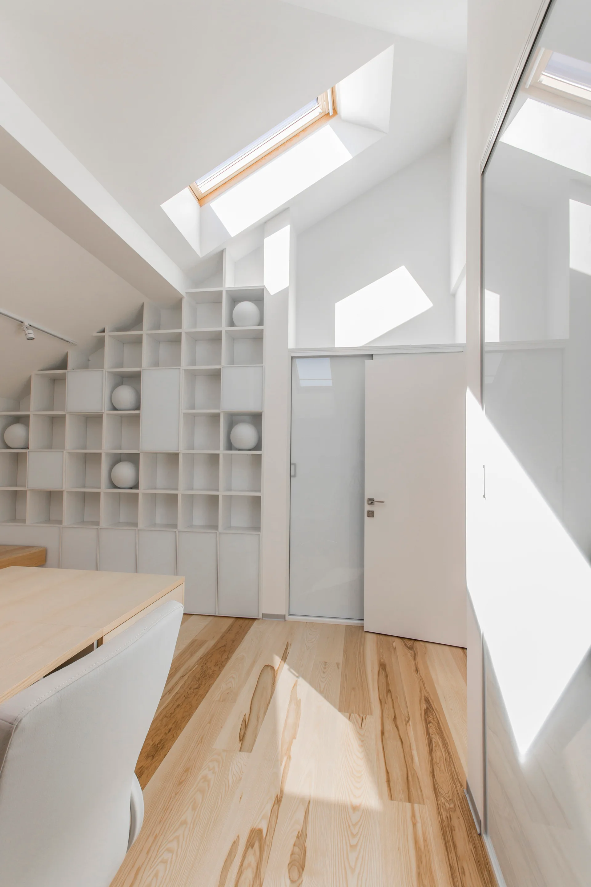 A minimalist attic room with a sloped ceiling, skylight, white custom grid shelving, and light wood flooring.