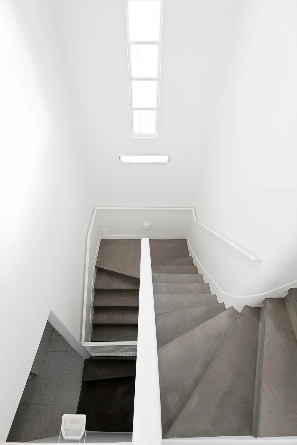 Looking down a winding concrete staircase with white handrails and walls illuminated by a narrow overhead skylight.