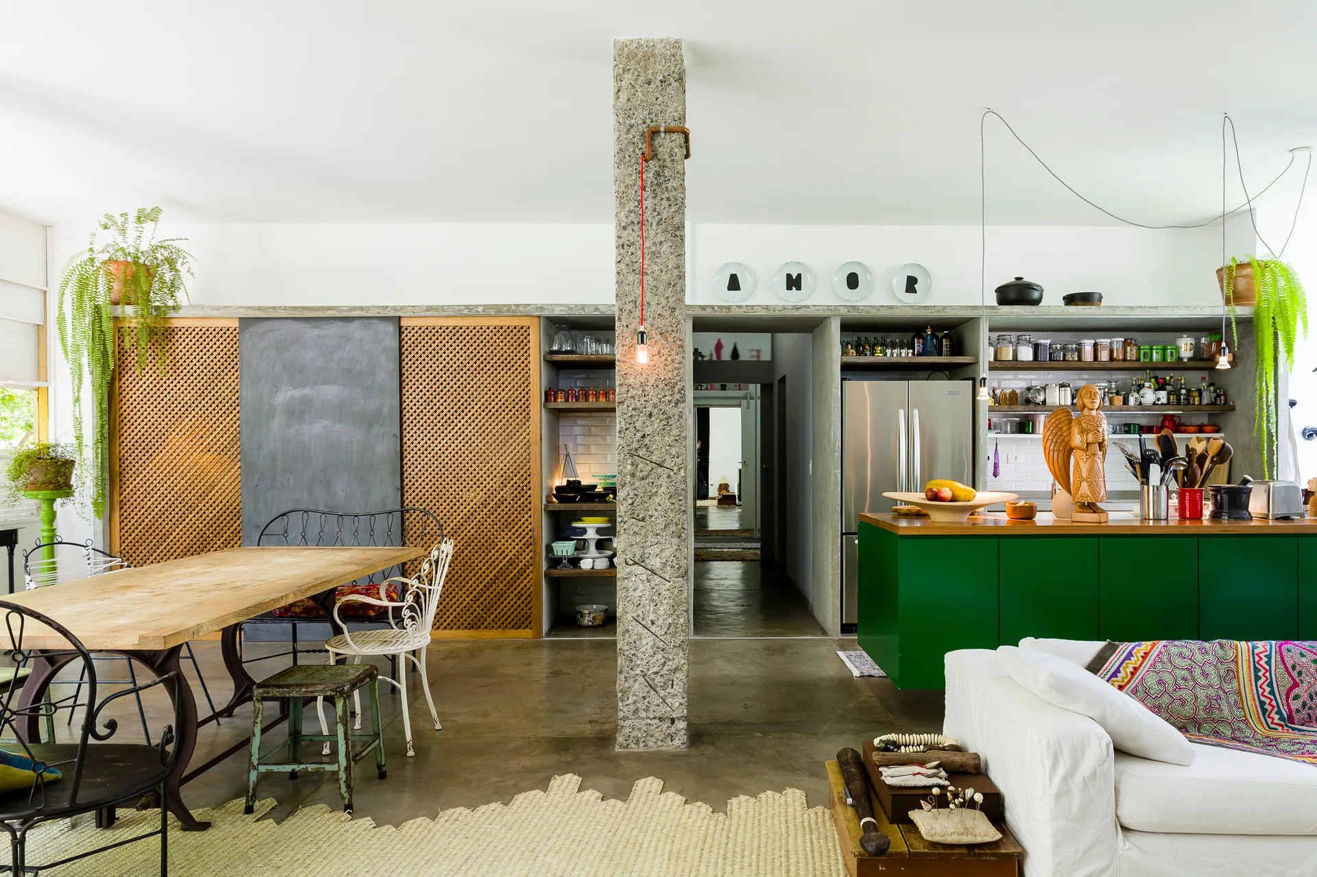 An open layout features a rough-hewn central stone column, a bold green kitchen island with wood countertop, and a dining table with mismatched chairs.