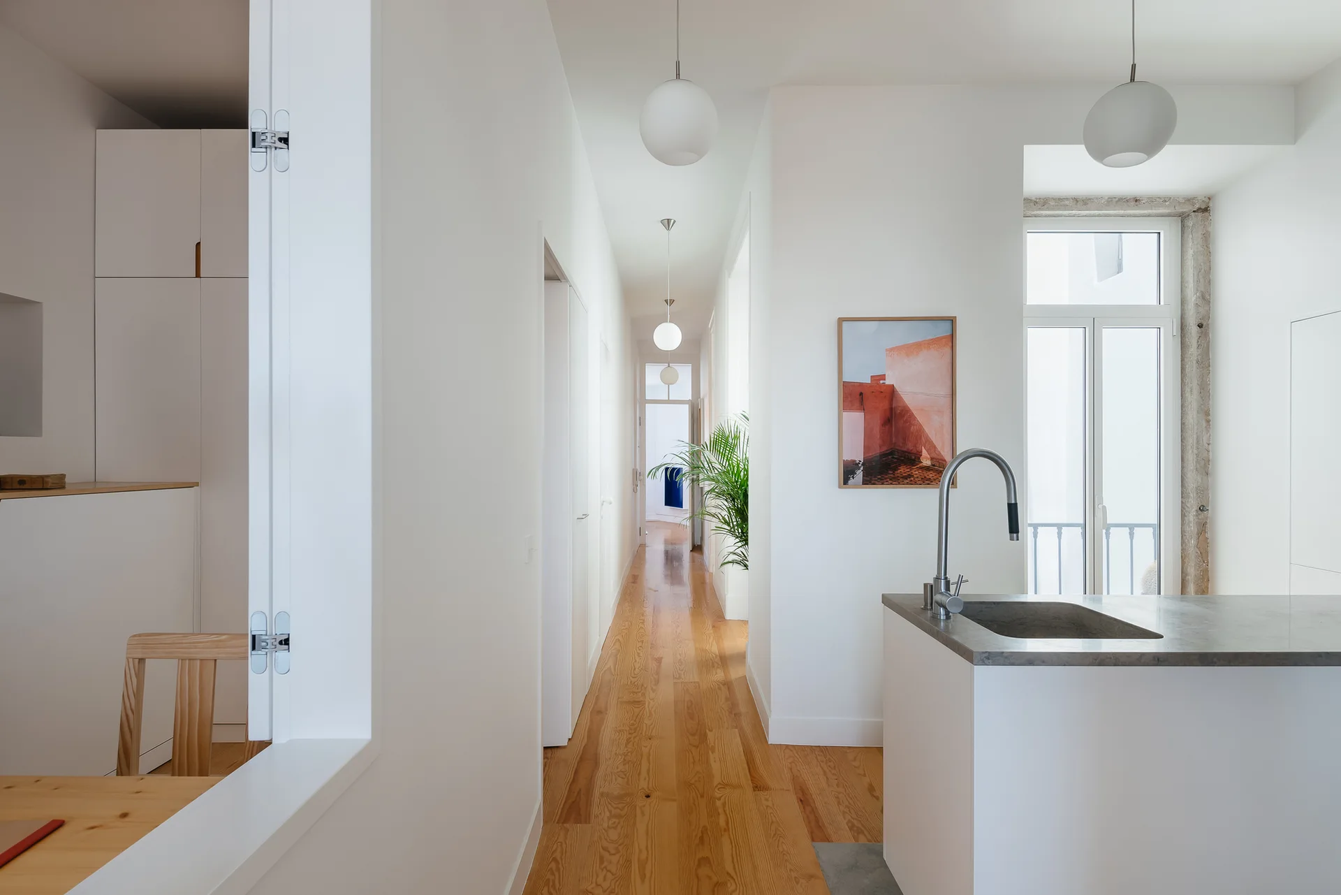 A bright minimalist hallway features light wood floors, globe pendant lights, and a view of a kitchen counter with a sink.