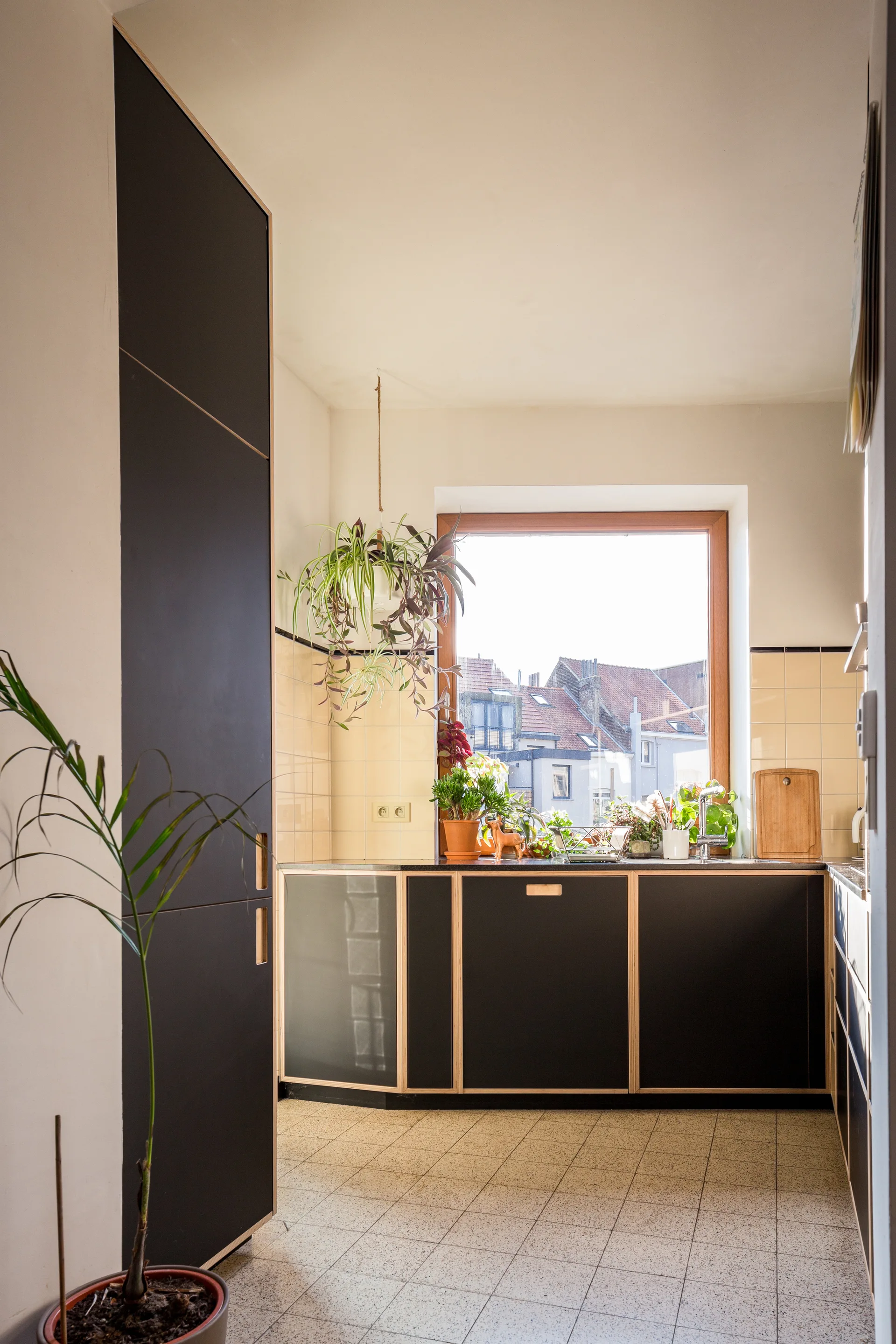 A residential kitchen featuring matte black cabinets with light plywood edges, a cream tiled backsplash, and a large wood-framed window.