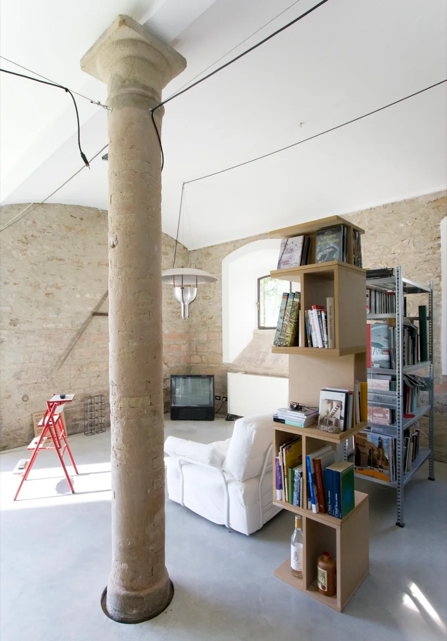 A living room with a prominent central stone column, exposed stone walls, a modular wooden bookshelf, and a white armchair on a grey floor.