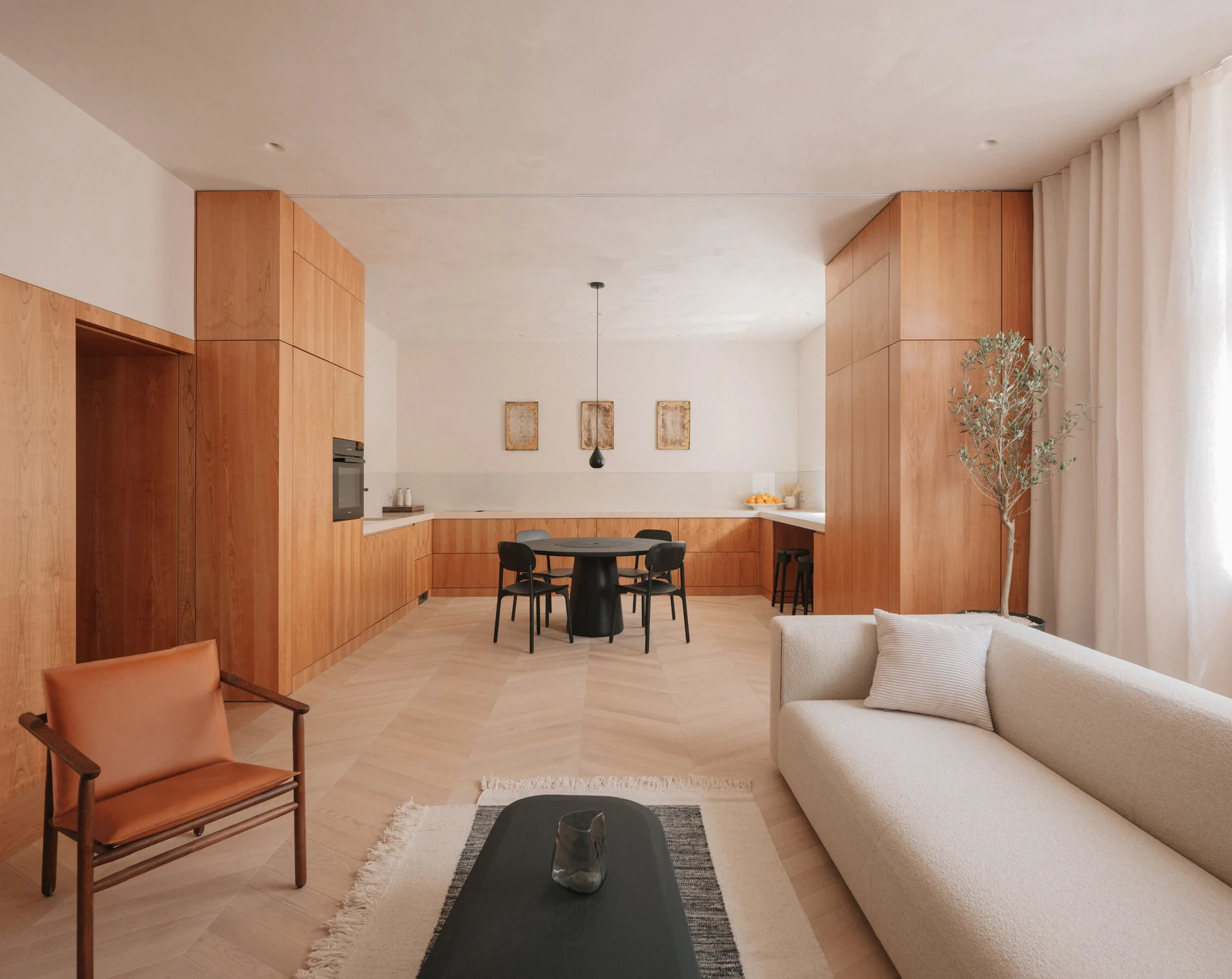 An open-plan living and dining area featuring a cream sofa, a leather armchair, herringbone wood flooring, and flat-panel wood cabinetry.