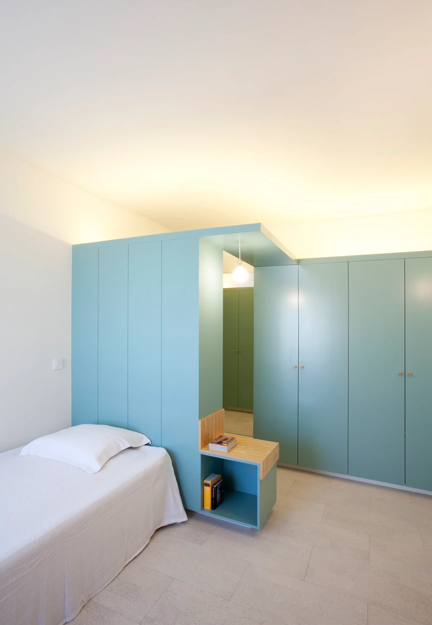 A minimalist bedroom featuring a single bed next to a large blue built-in wardrobe partition with an integrated wooden nightstand.