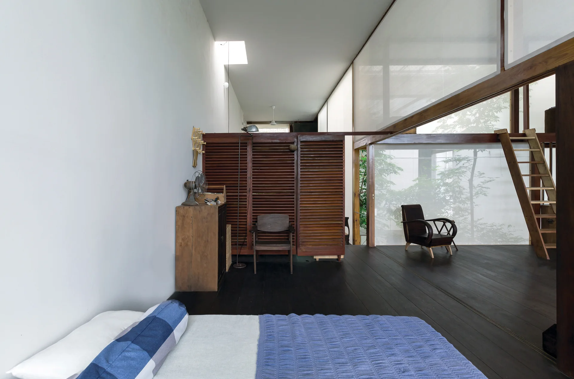 A minimalist bedroom featuring dark wood flooring, a central wooden louvered room divider, and large translucent window screens.