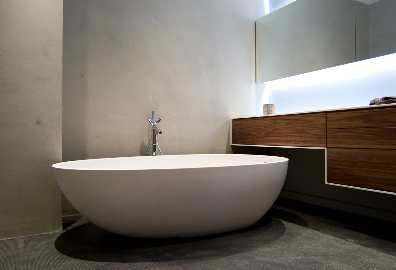 A white oval freestanding bathtub sits against a textured grey concrete wall next to a wall-mounted wooden vanity with an illuminated mirror.