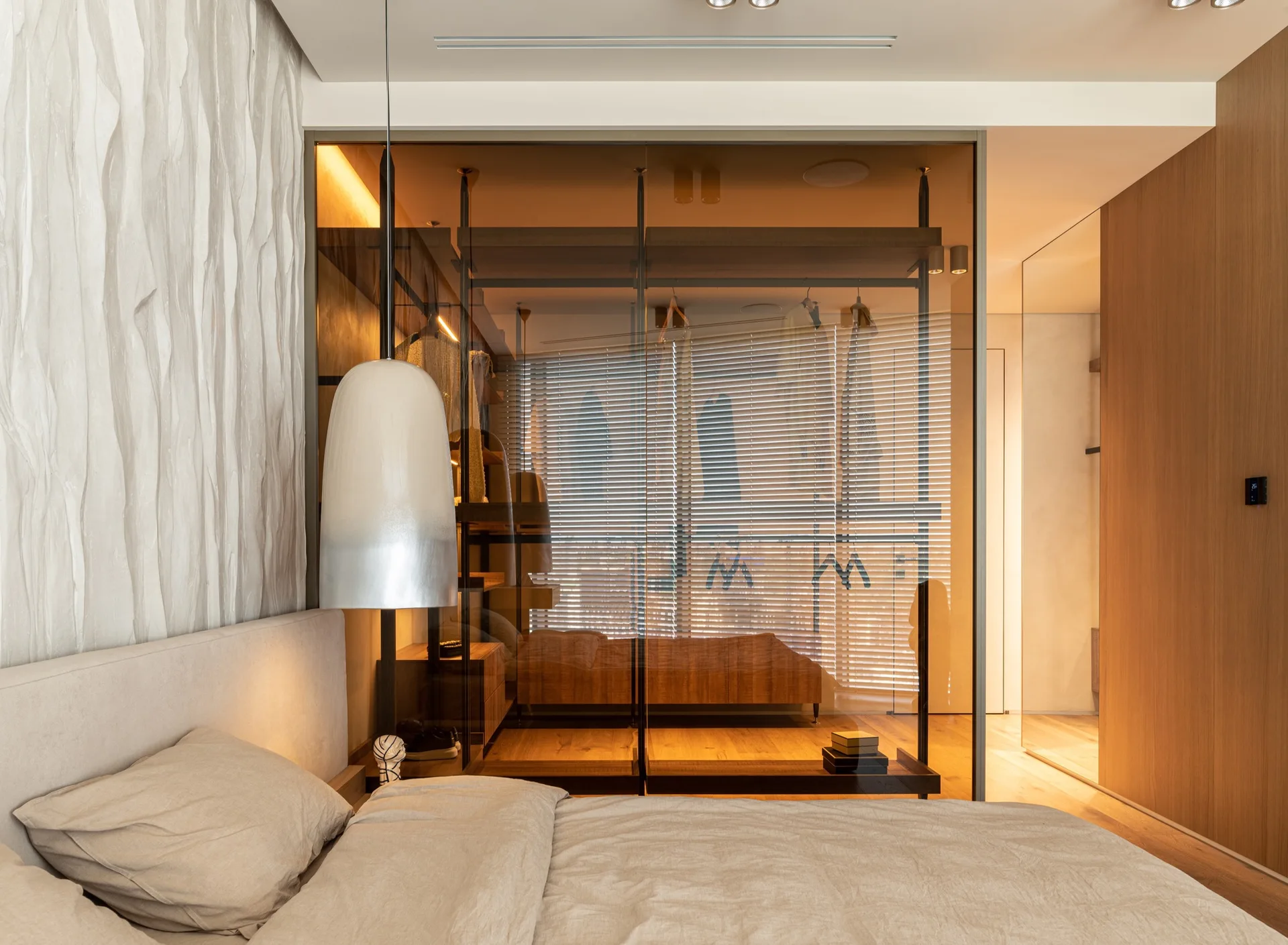 A modern bedroom features a bed with beige bedding, a wavy textured accent wall, and a tinted glass closet with warm interior lighting.