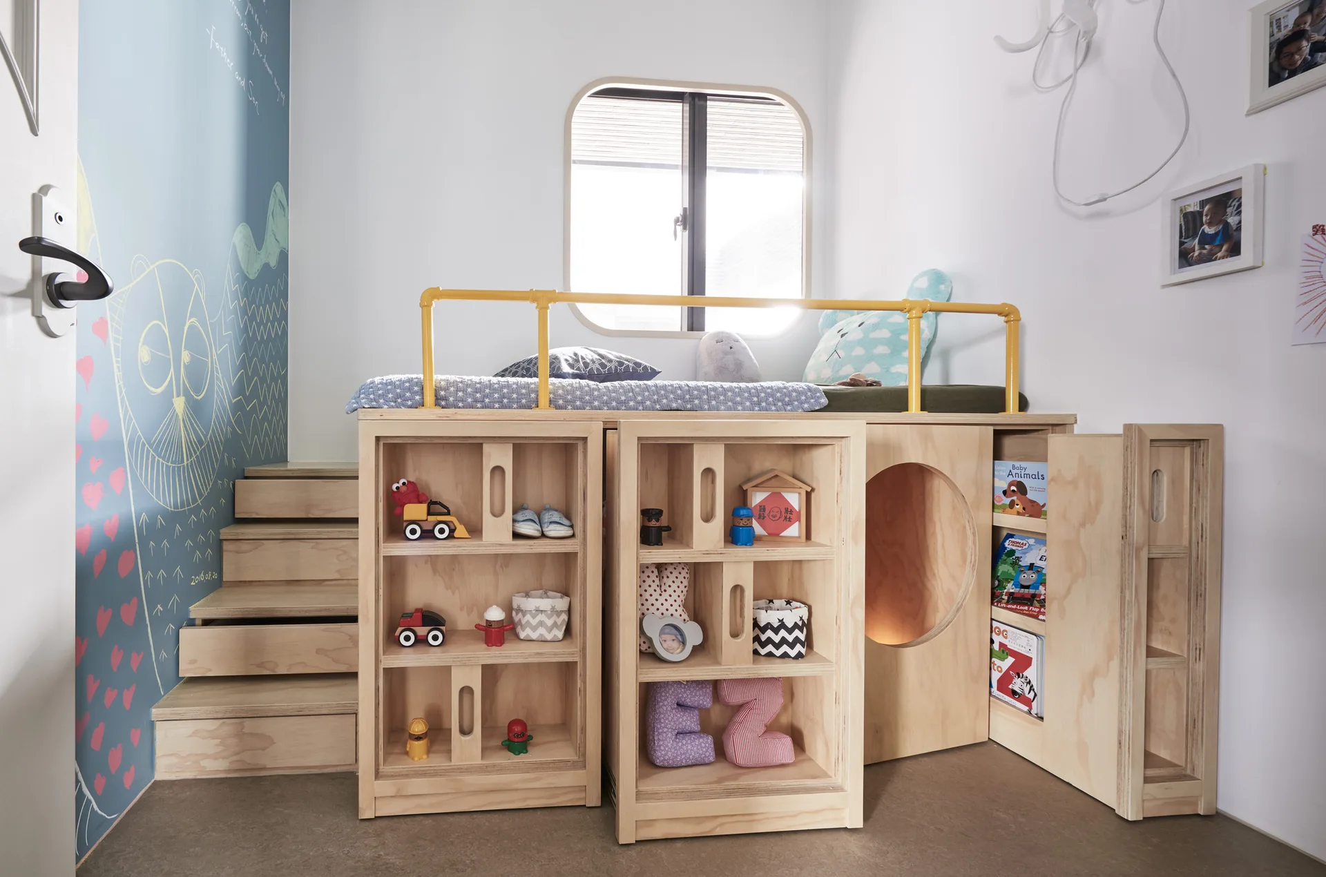 A children's room features a custom plywood loft bed with integrated wooden storage shelves, stairs, and a bright yellow metal railing.