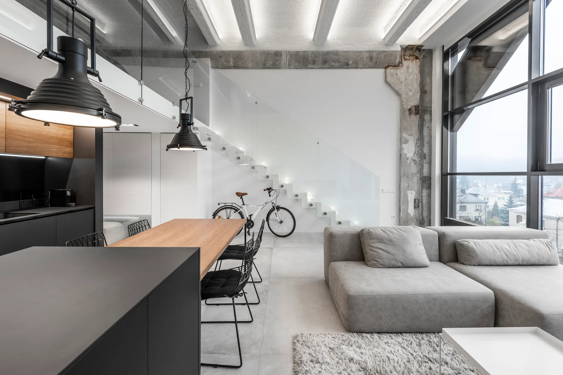 An open-plan industrial loft featuring a grey sectional sofa, a wood-topped dining table with black wire chairs, and a floating white staircase.