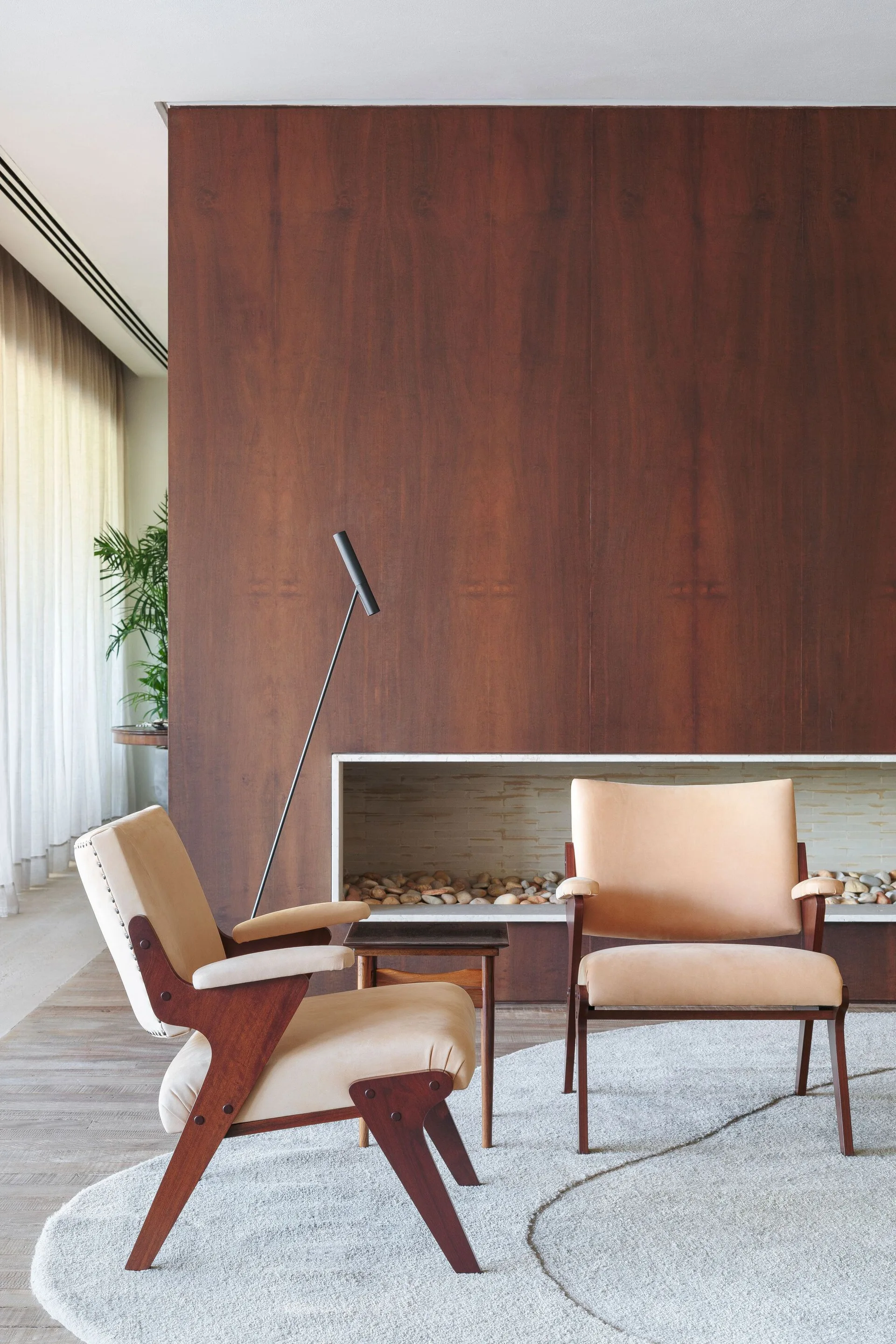 Two tan mid-century armchairs sit on a light gray oval rug in front of a dark wood-paneled wall with a low linear fireplace filled with pebbles.