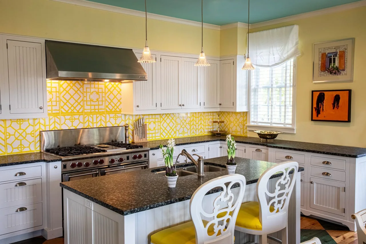 A kitchen with white beadboard cabinets, a yellow geometric tile backsplash, dark granite countertops, and a painted teal ceiling.