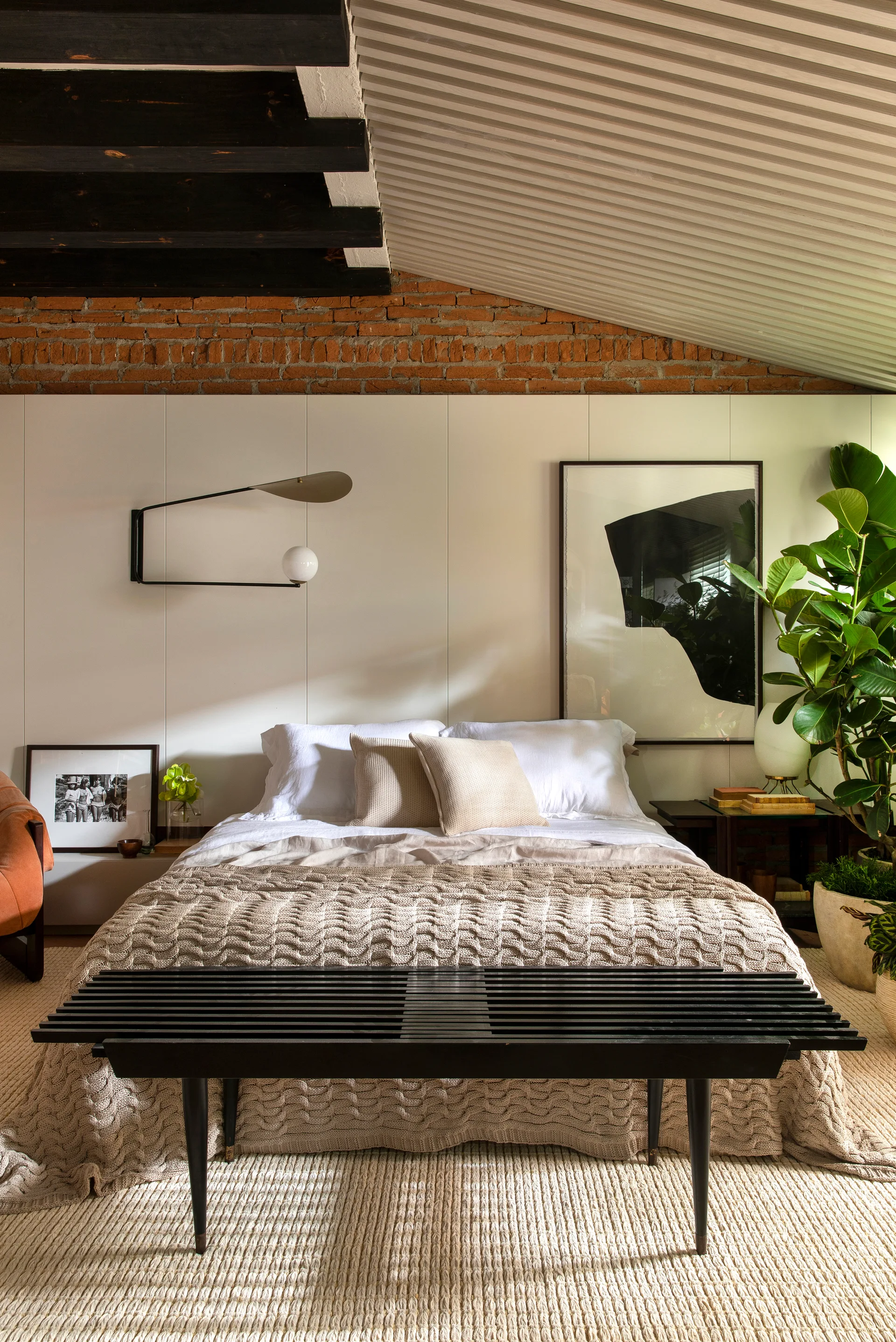 A cozy bedroom featuring a black slatted bench at the foot of a bed, an exposed brick wall section, and dark rustic ceiling beams.