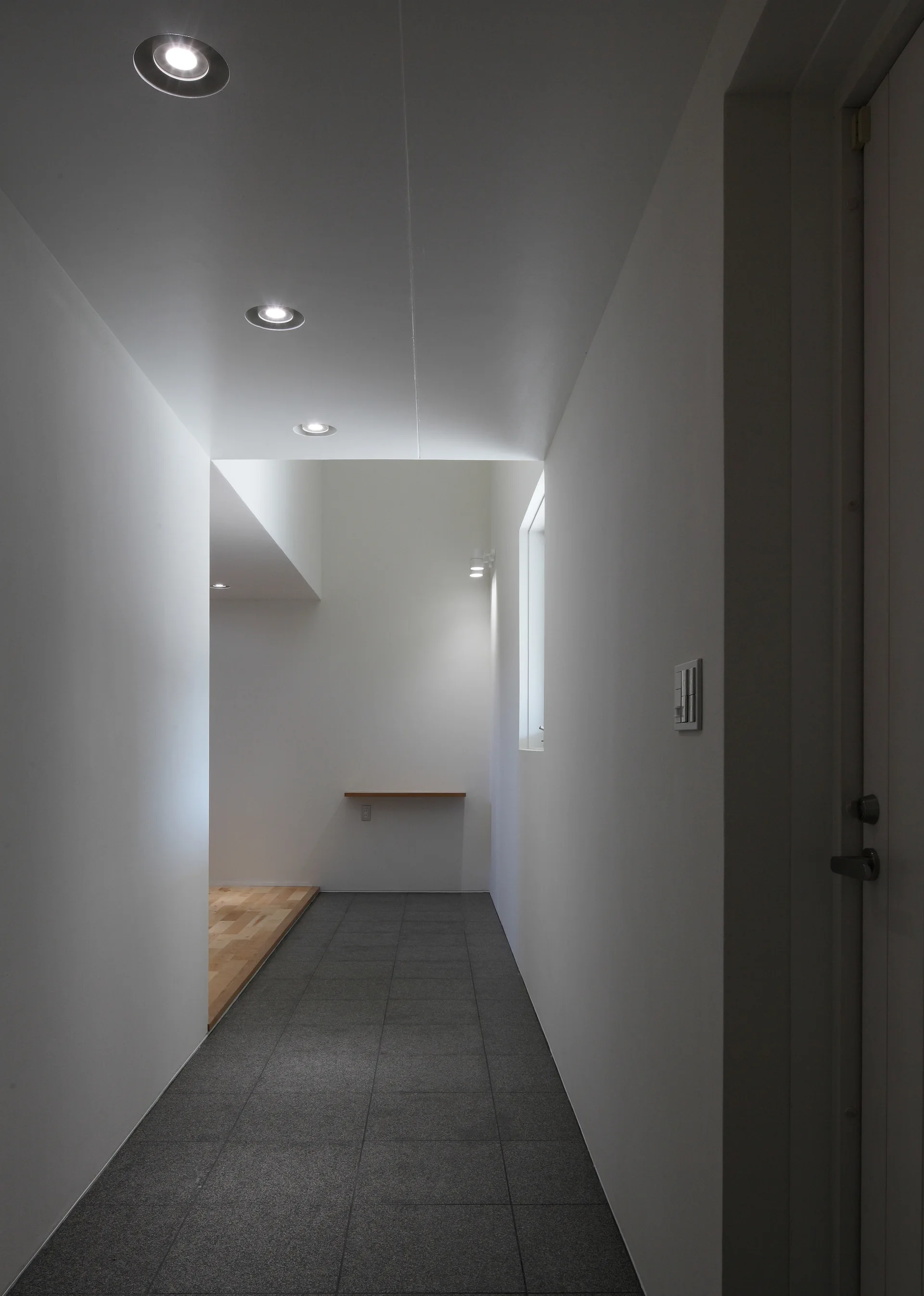 A narrow white hallway with grey tiled flooring that transitions to light wood, lit by round recessed ceiling downlights.