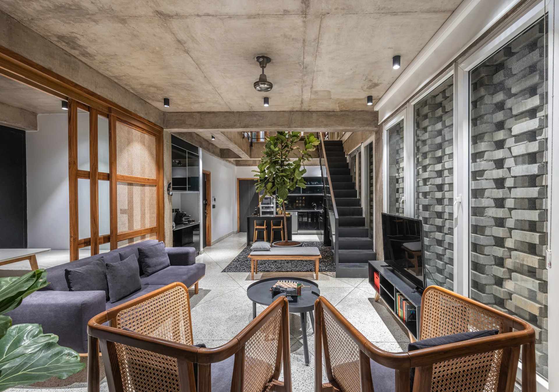 An industrial living room features a concrete ceiling, terrazzo floor, a grey sofa, and rattan-paneled armchairs near a tall fiddle-leaf fig tree.