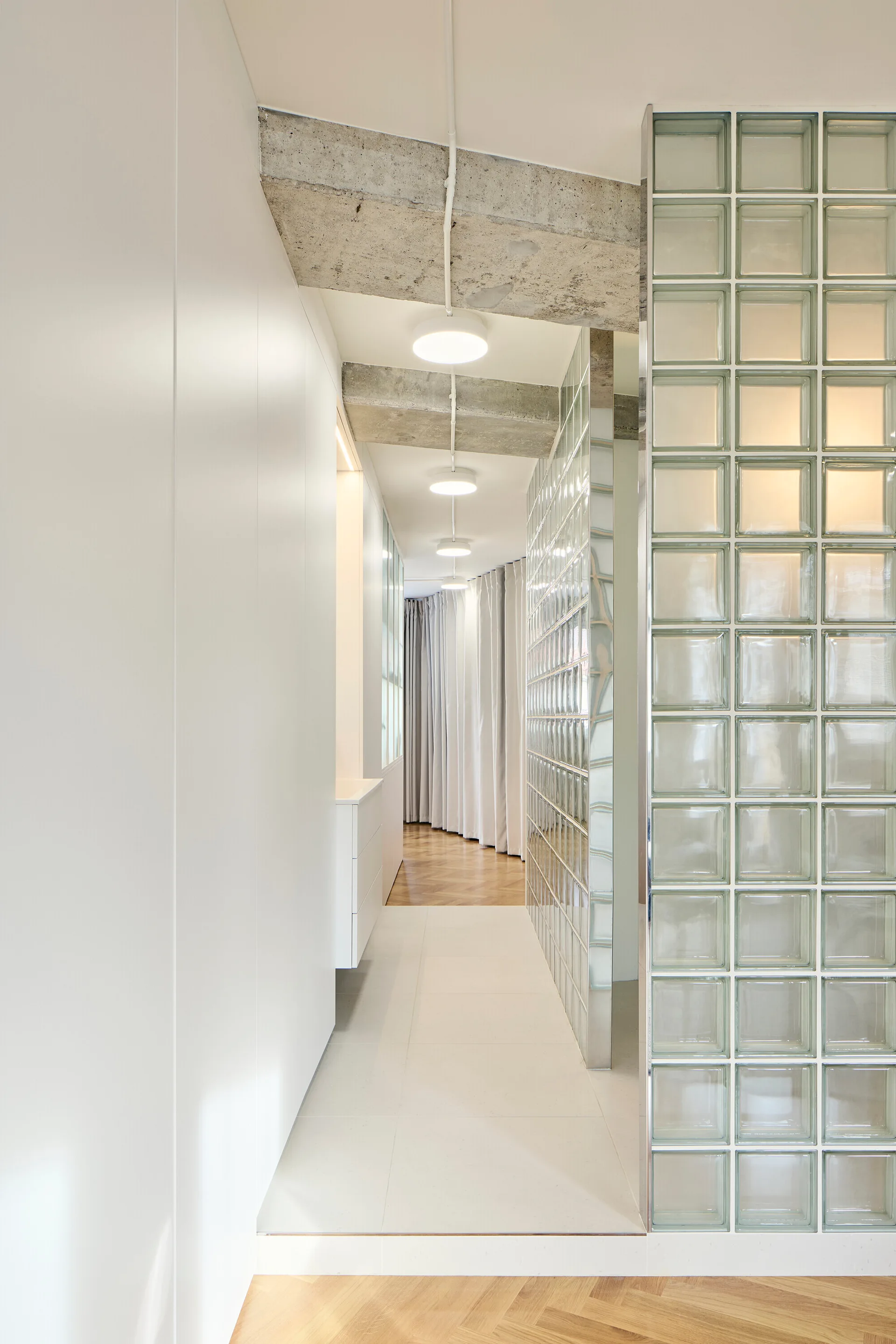 A clean white hallway features a translucent glass block partition wall, raw concrete ceiling beams, and integrated white storage cabinets.