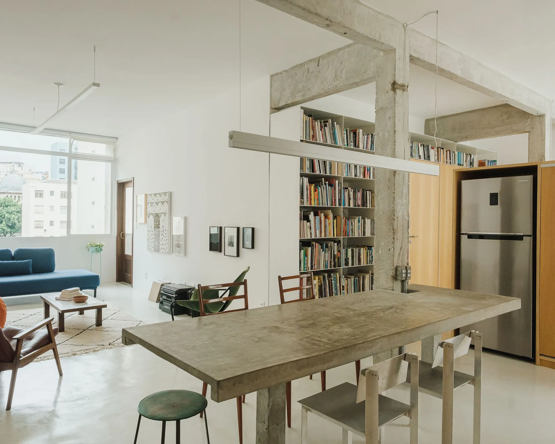 An open-plan apartment featuring a raw concrete dining table, exposed concrete columns, a tall white bookcase, and a blue sofa.