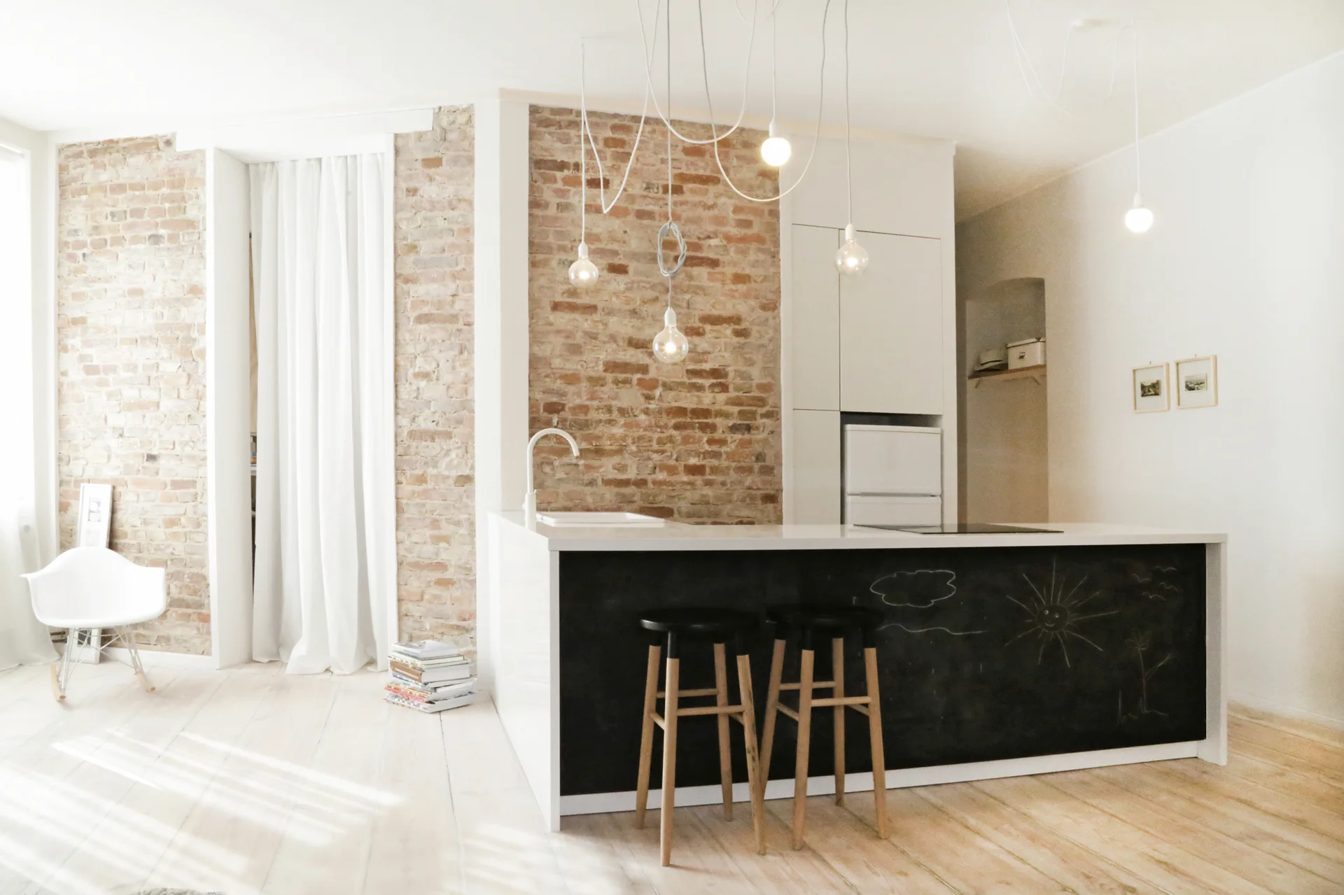 A minimalist white kitchen featuring a central island with a chalkboard front, wooden bar stools, and an exposed brick accent wall.