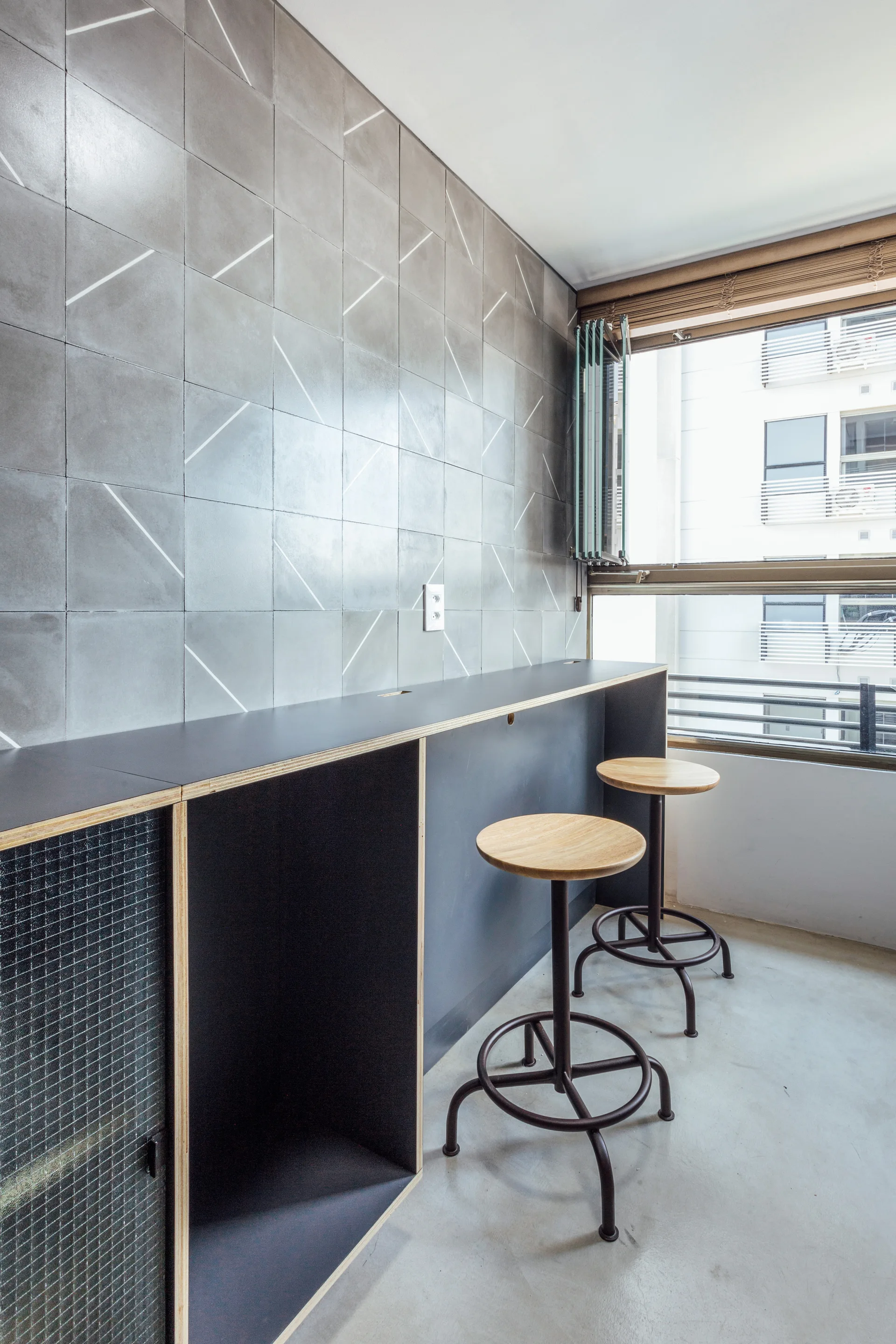 A minimalist breakfast bar features a dark plywood-edged counter, wooden-topped stools, and a feature wall with grey geometric tiles.