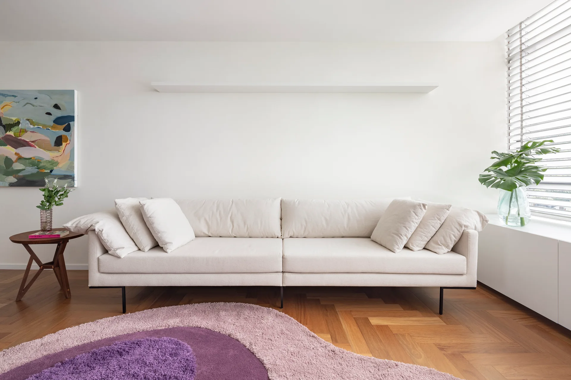 A cream sofa sits on herringbone wood floors next to a circular purple rug in a bright, white-walled living room with a floating shelf.
