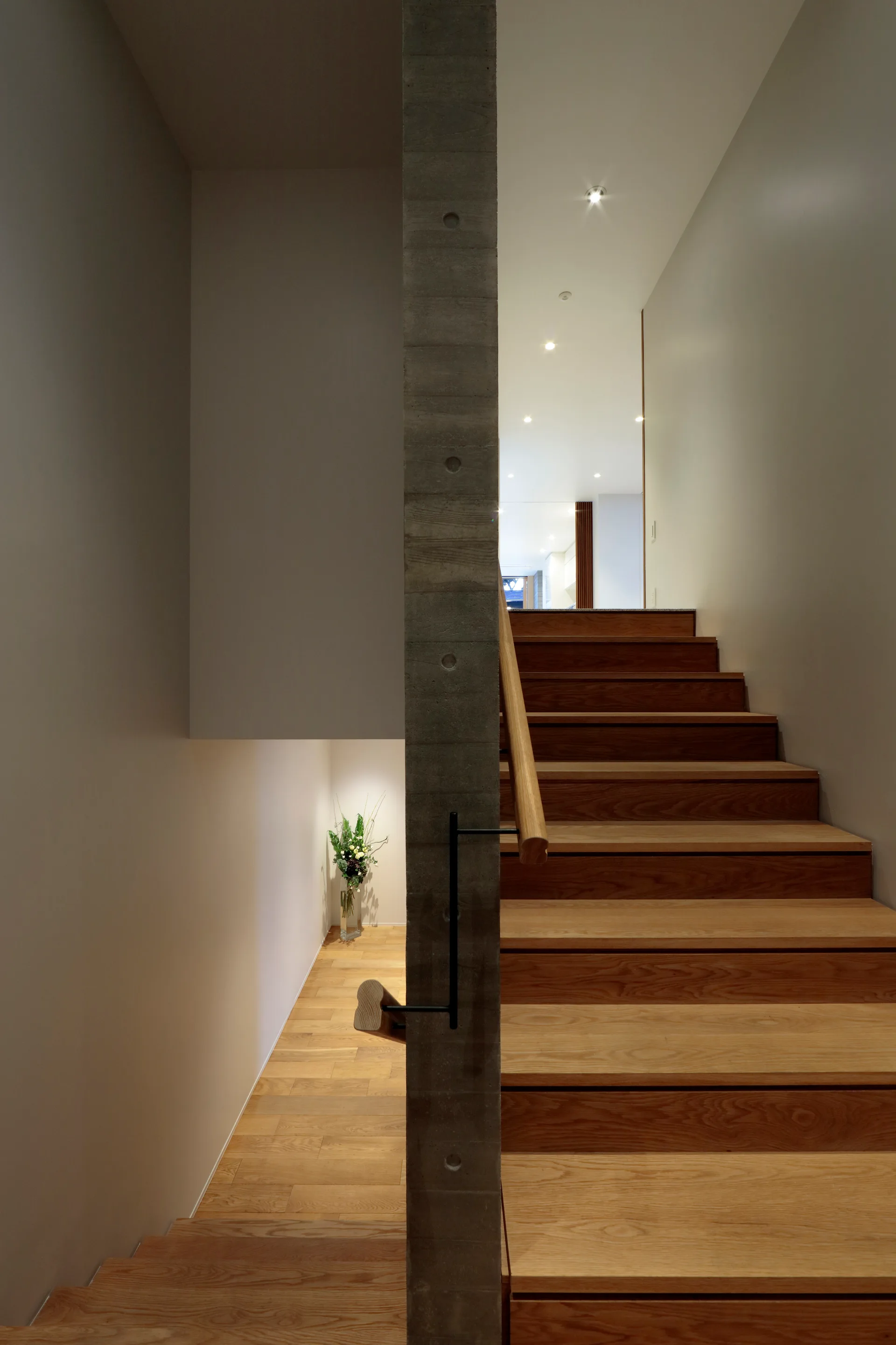A modern staircase with light wood steps features a vertical raw concrete column and a wooden handrail with black metal hardware.