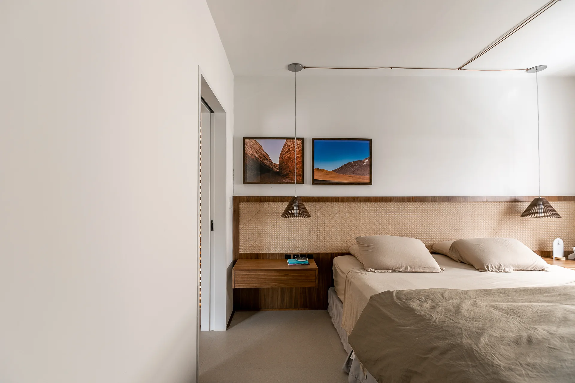 A minimalist bedroom featuring a bed with beige bedding, a wide woven wicker and wood headboard, a floating nightstand, and two framed landscape photos.