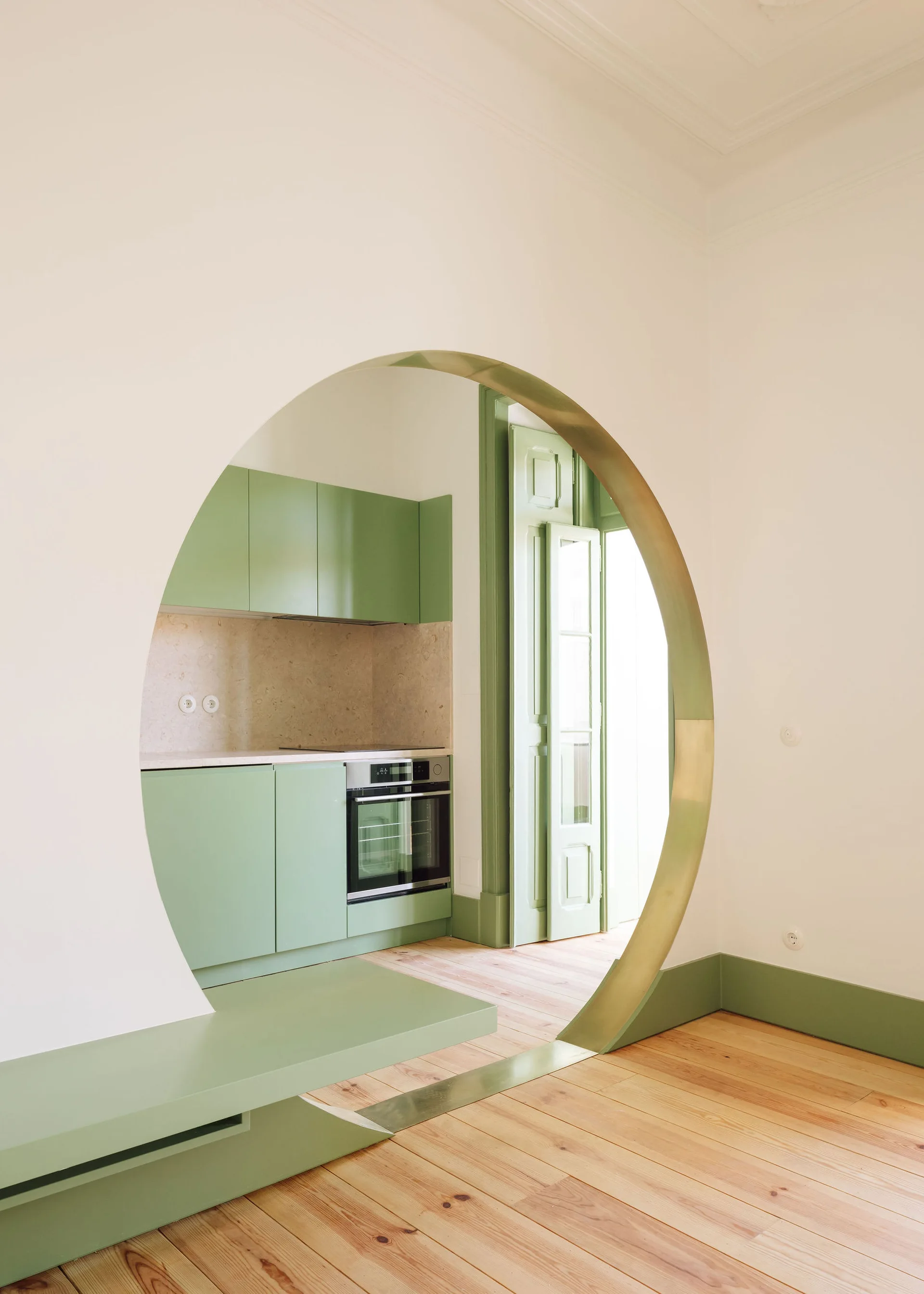 A large circular archway with metallic brass trim frames a sage green kitchen with matching cabinetry, built-in oven, and light wood flooring.