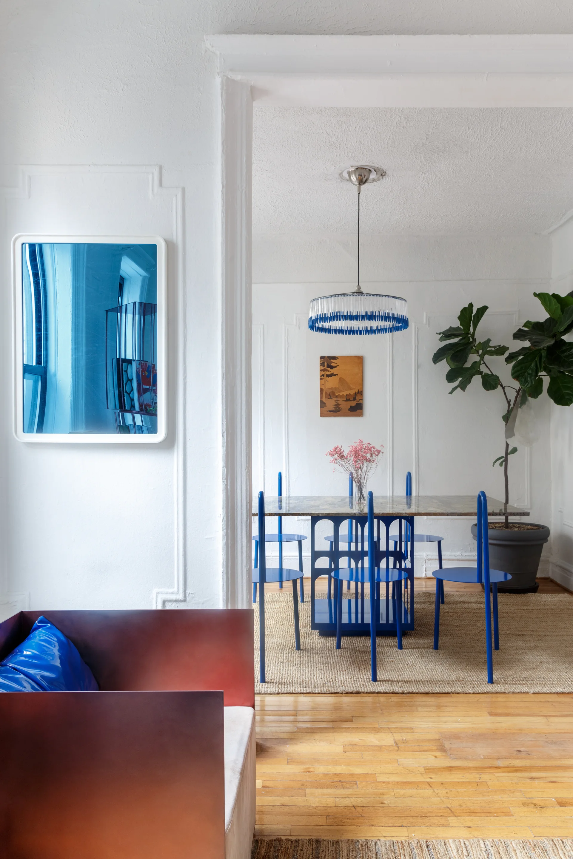 A dining room seen through an archway features cobalt blue metal chairs, a stone-topped table, a woven rug, and white paneled walls.