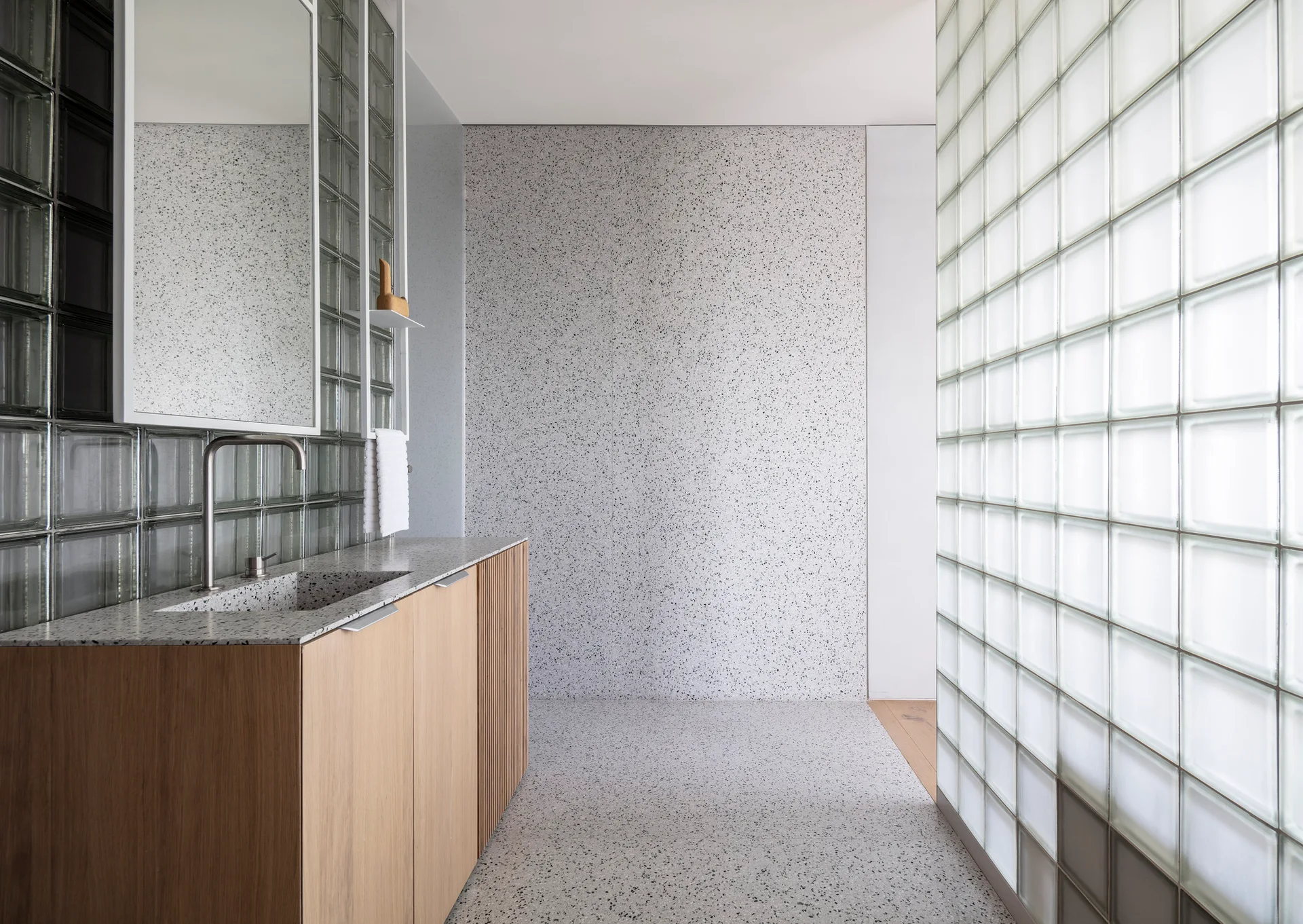 A modern bathroom features a light wood vanity with an integrated speckled sink, a glass block partition wall, and matching terrazzo floors.