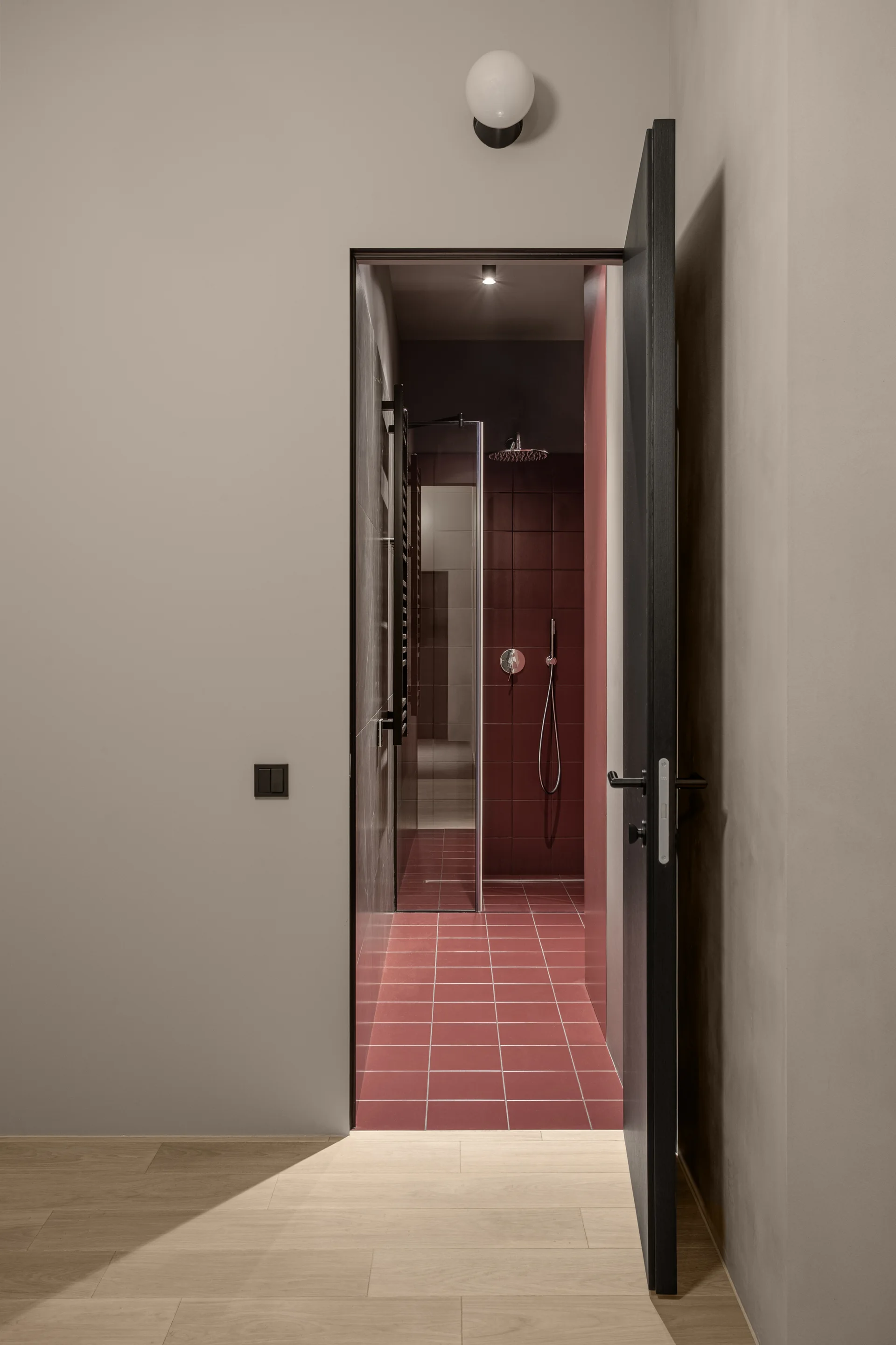 A view from a beige hallway through a dark open door reveals a bathroom lined with matte red square ceramic tiles and black fixtures.