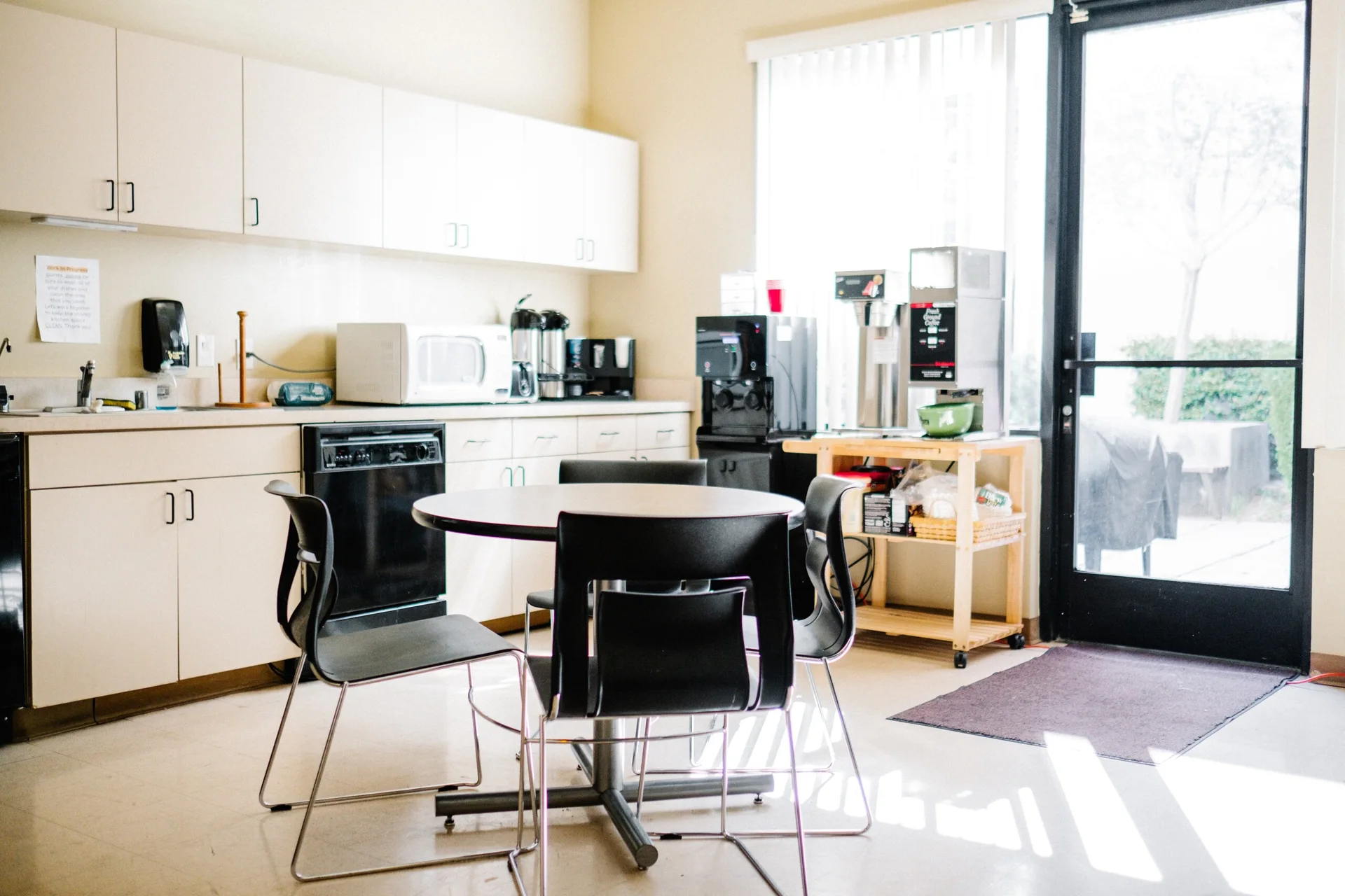 A compact office kitchenette features off-white upper and lower cabinets, a round table with four black chairs, and a wooden cart beside a glass door.