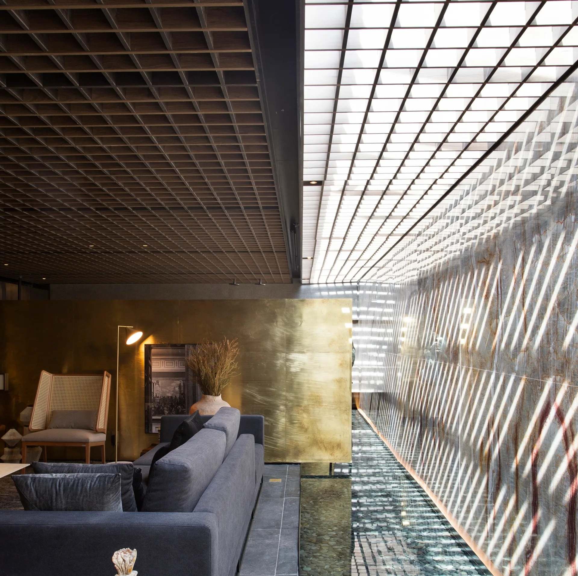 A contemporary living room featuring a dark grey sofa, a coffered wood ceiling grid, and an indoor water channel running alongside a stone wall.