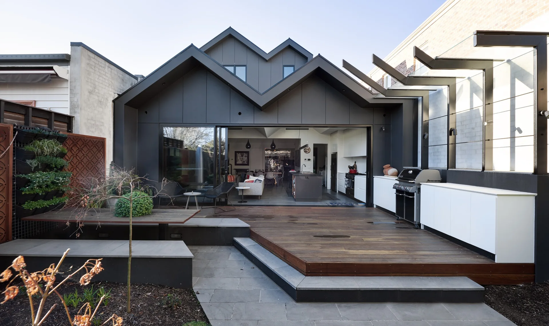 A modern wood-plank deck features a white outdoor kitchen and barbecue grill, framed by a charcoal-colored geometric home exterior with sliding glass doors.
