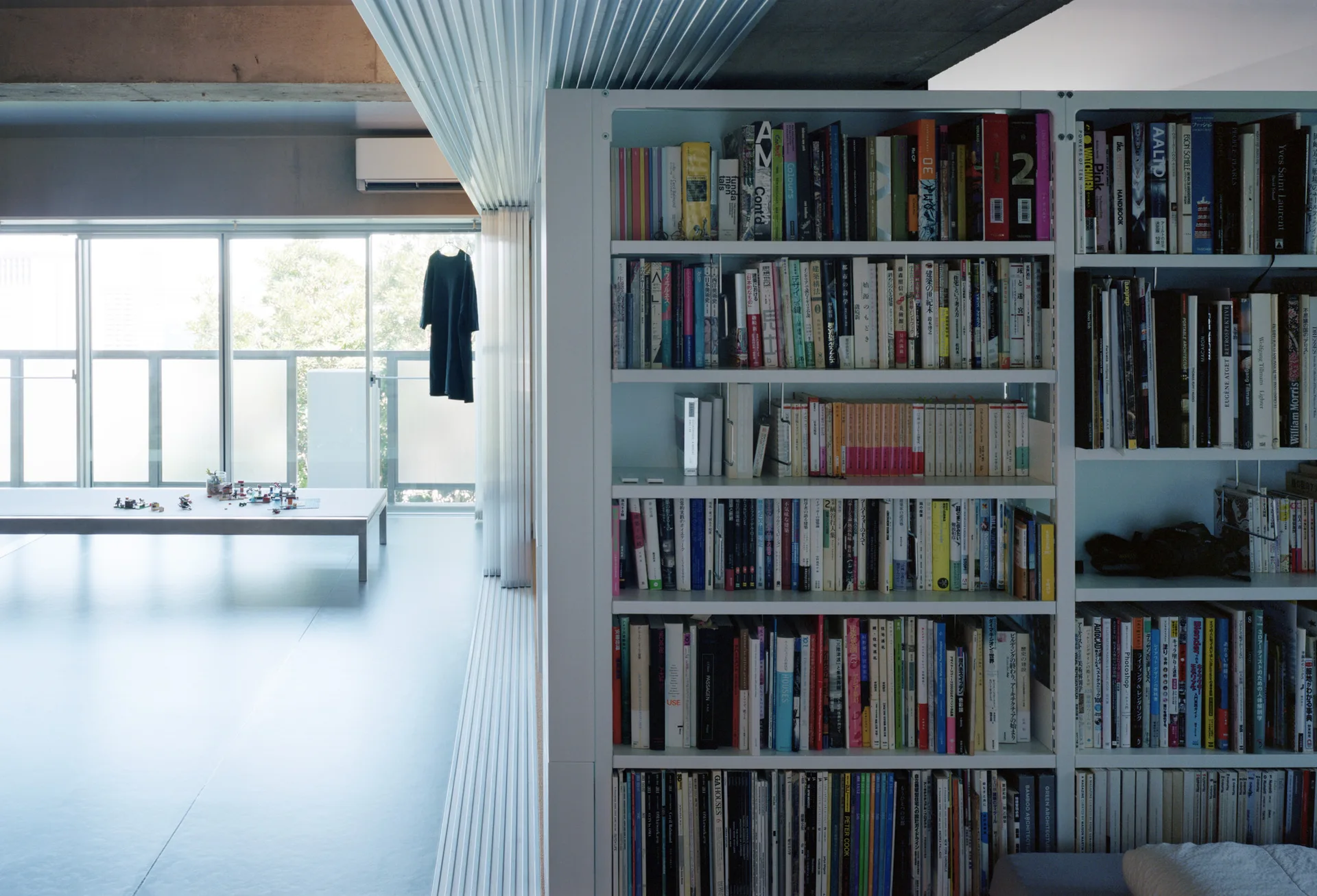 A massive white metal bookshelf filled with books stands next to a minimalist room with a low table, concrete ceiling, and large windows.