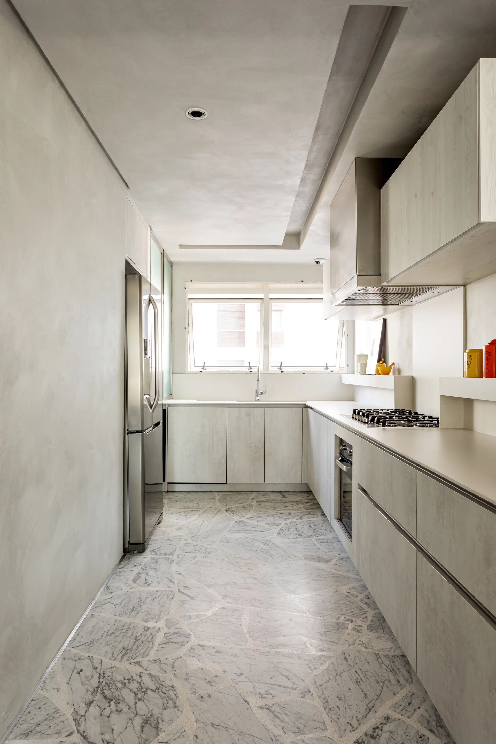 A minimalist galley kitchen features light grey flat-front cabinets, stainless steel appliances, and a unique floor made of broken marble pieces.