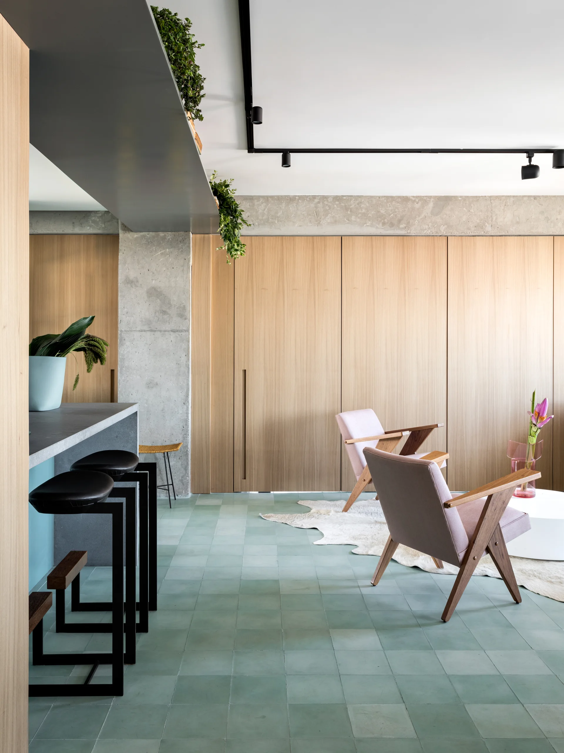 A modern space features pastel green floor tiles, floor-to-ceiling wood paneling, an exposed concrete column, and pink mid-century armchairs.