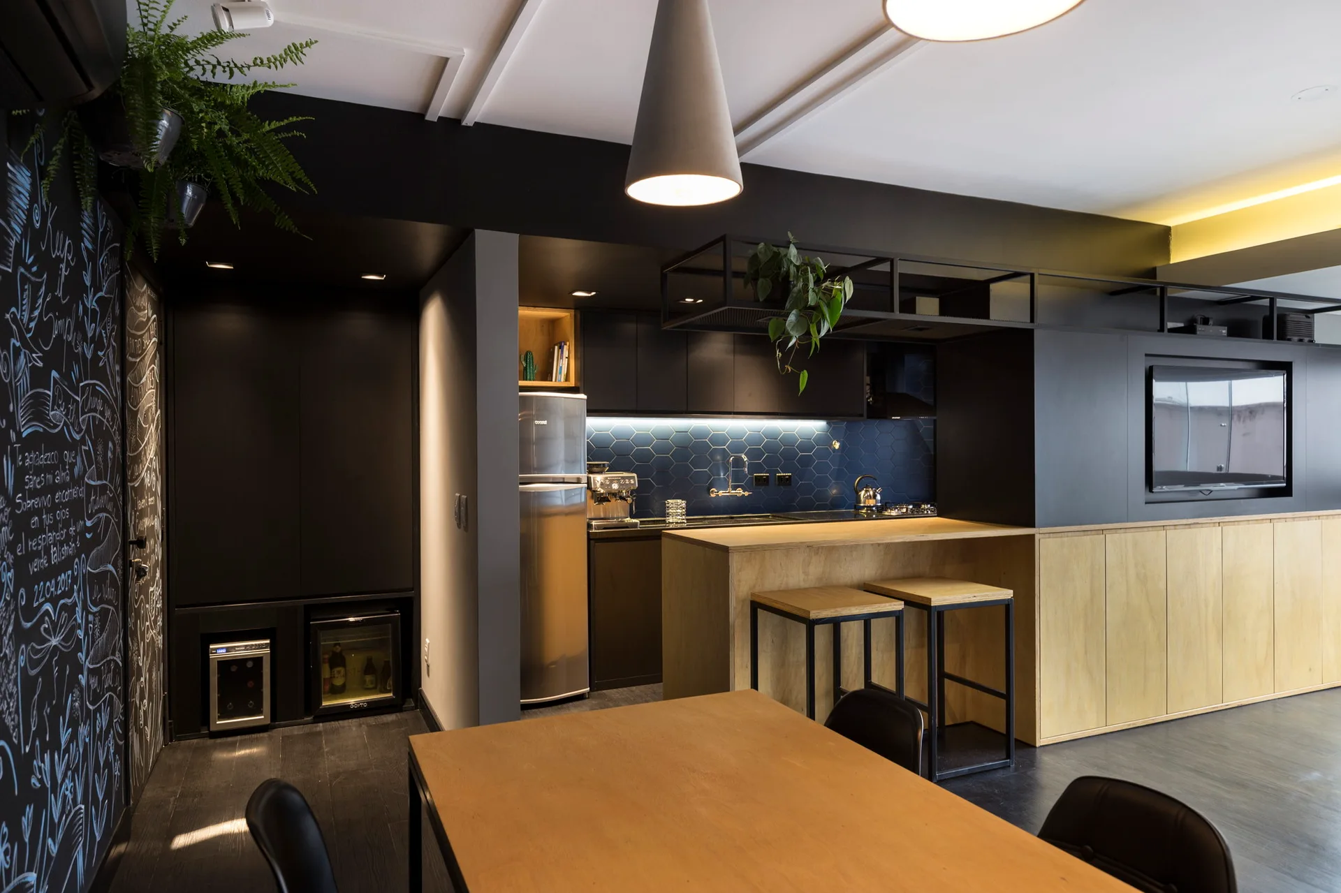 A modern kitchen features matte black cabinetry, a blue hexagon tile backsplash, a wood-toned island with stools, and a black chalkboard wall.