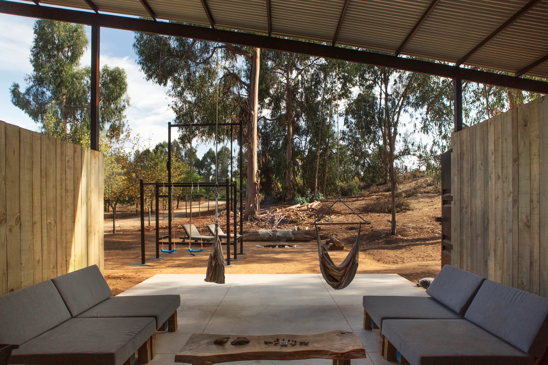 A covered concrete patio features grey modular sofas, a raw-edge wood coffee table, and hanging hammocks overlooking a rustic outdoor landscape.