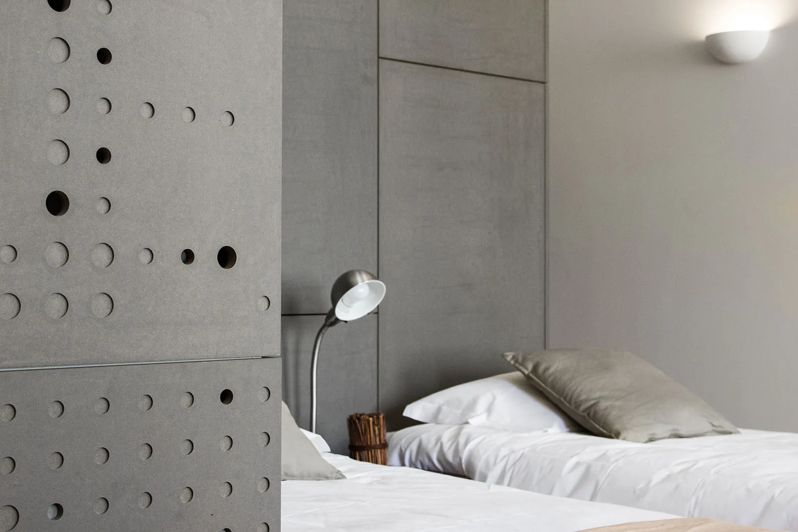 A minimalist bedroom featuring twin beds with white sheets in front of large grey wall panels and a perforated concrete screen.