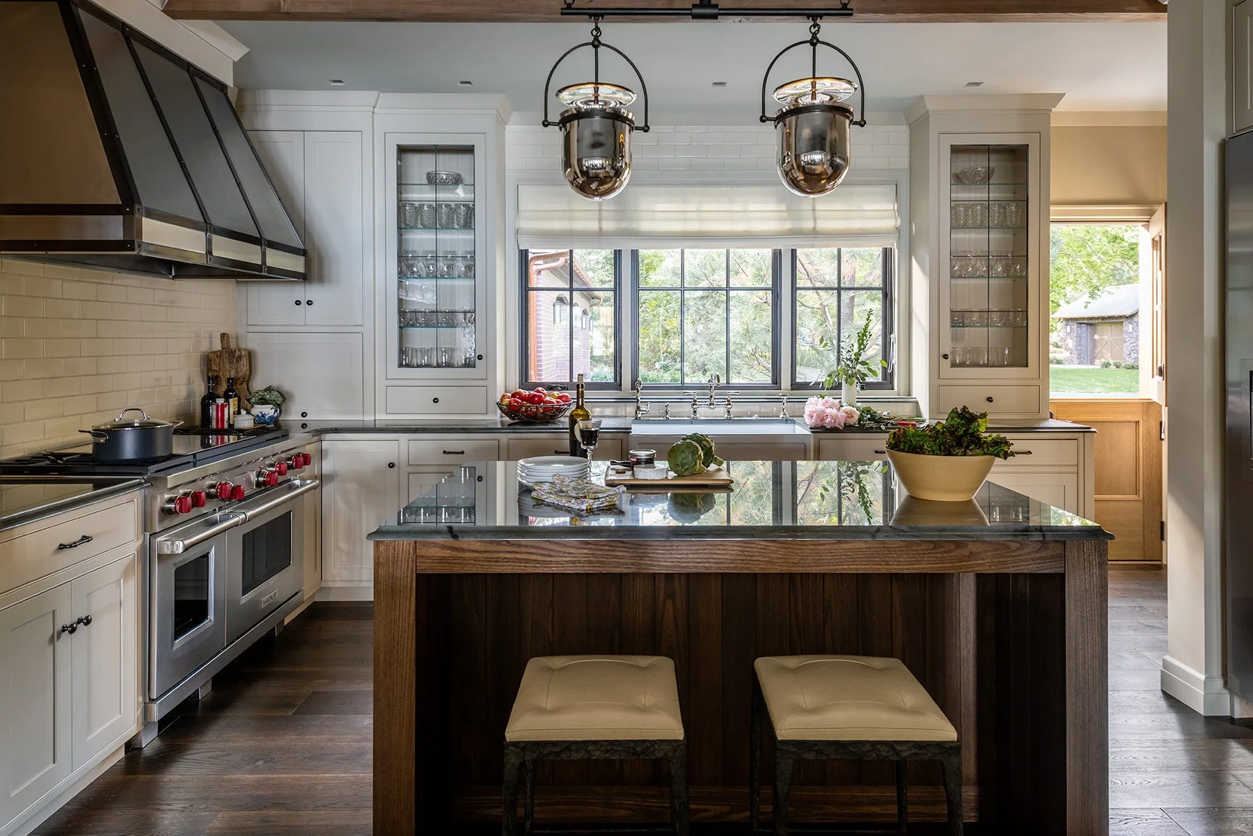 A residential kitchen features a dark wood island with stools, cream-colored cabinetry, a professional range under a metal hood, and two pendant lights.