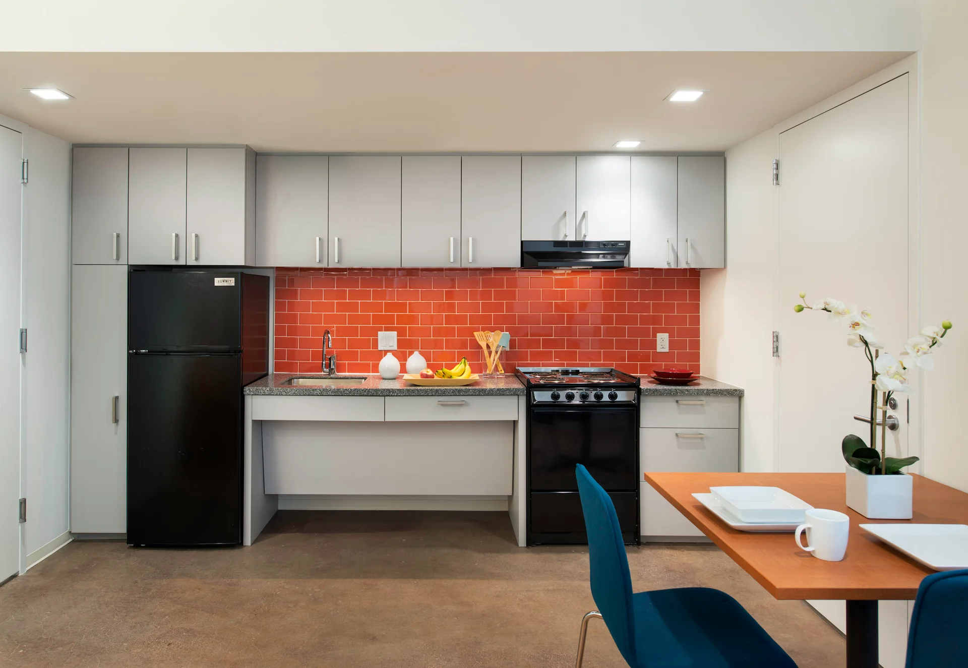 A compact kitchen features light grey cabinetry, a red subway tile backsplash, and a wooden dining table with blue chairs.
