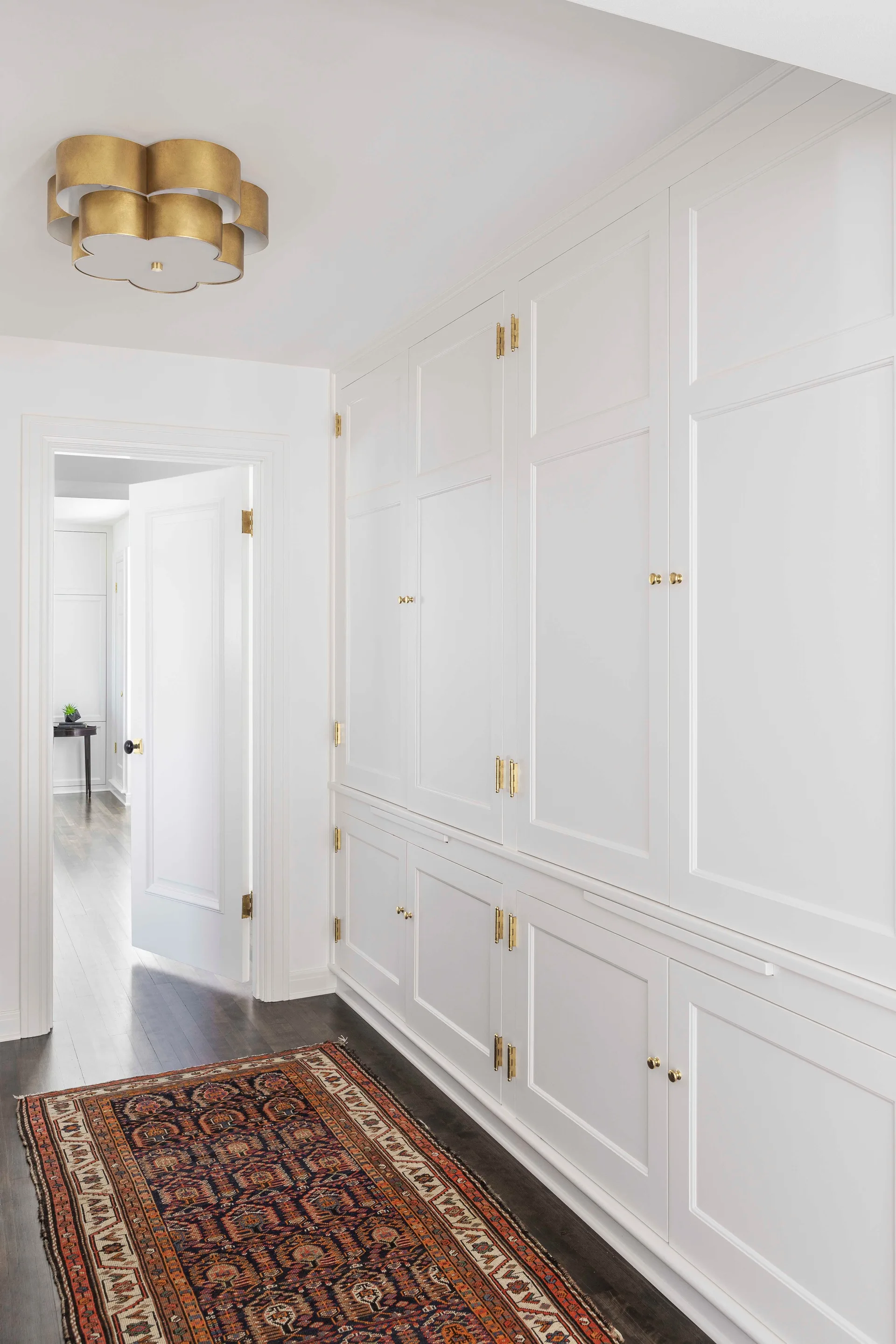A white hallway features extensive built-in cabinetry with brass hardware, a patterned runner rug on dark wood floors, and a gold ceiling light.