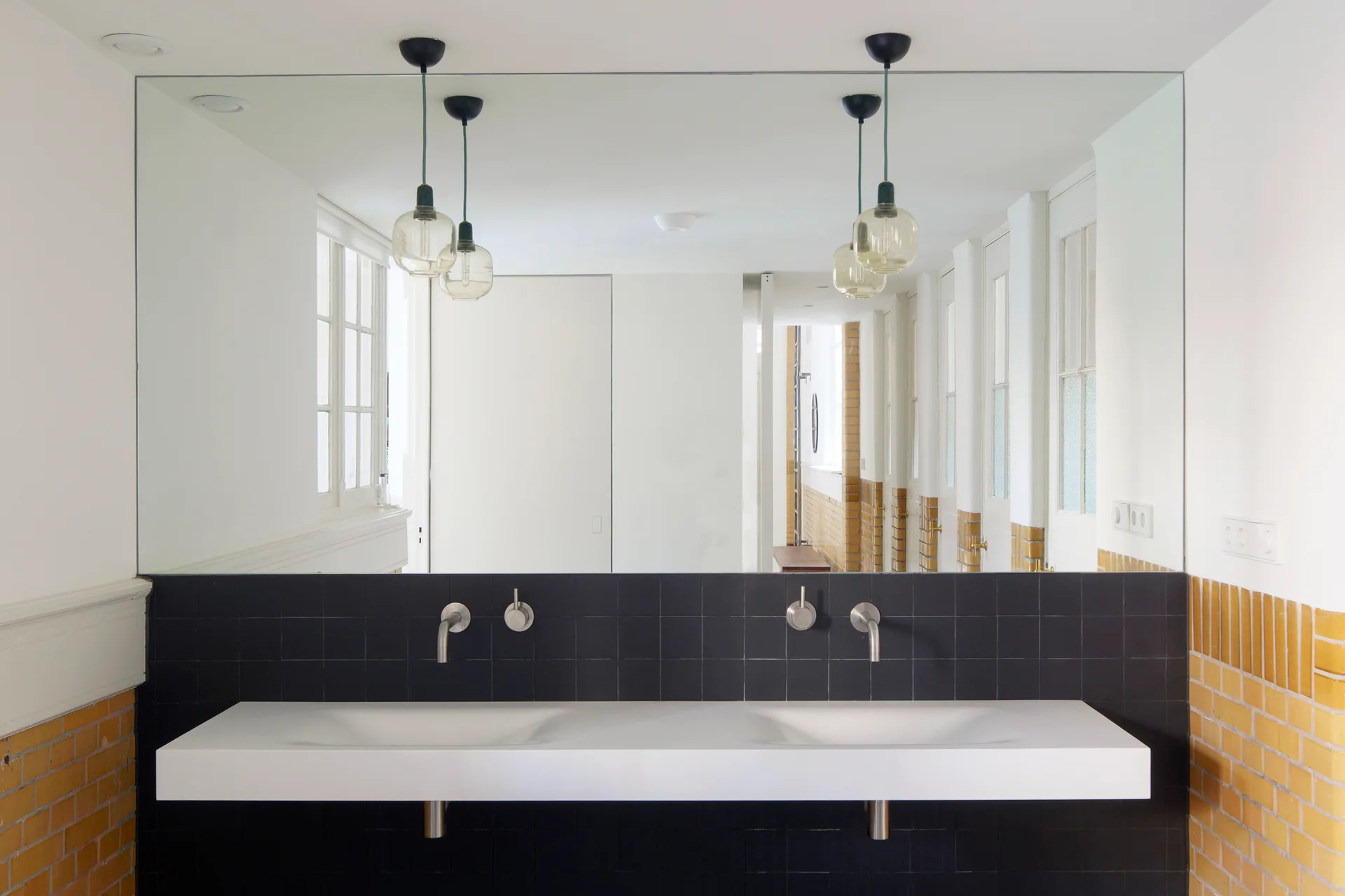 A minimalist double-sink vanity with wall-mounted faucets is set against a black tiled wall beneath a large frameless mirror and glass pendant lights.